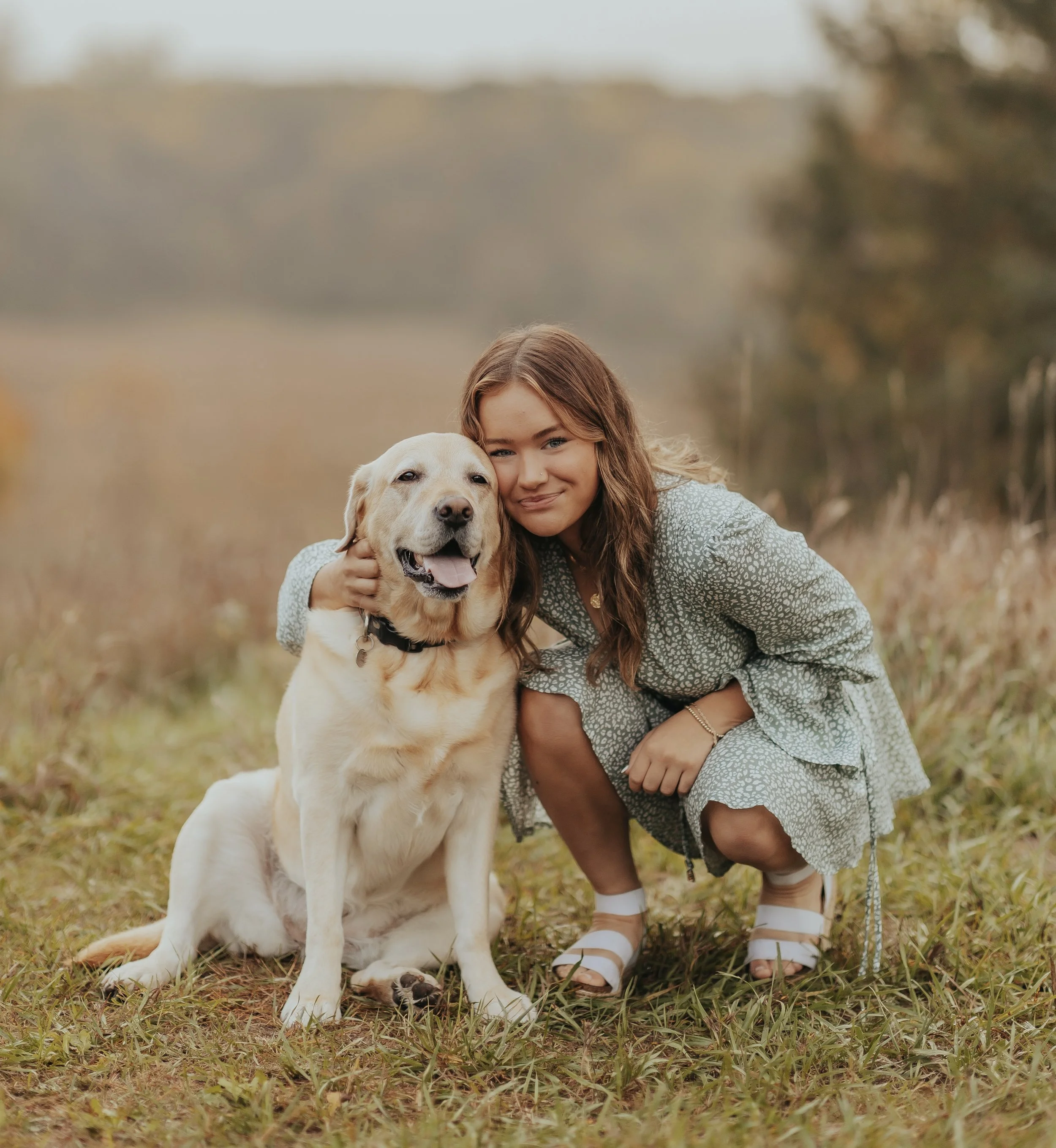 A young woman crouching next to a yellow Labrador retriever in an outdoor field during fall, with both smiling and looking at the camera.