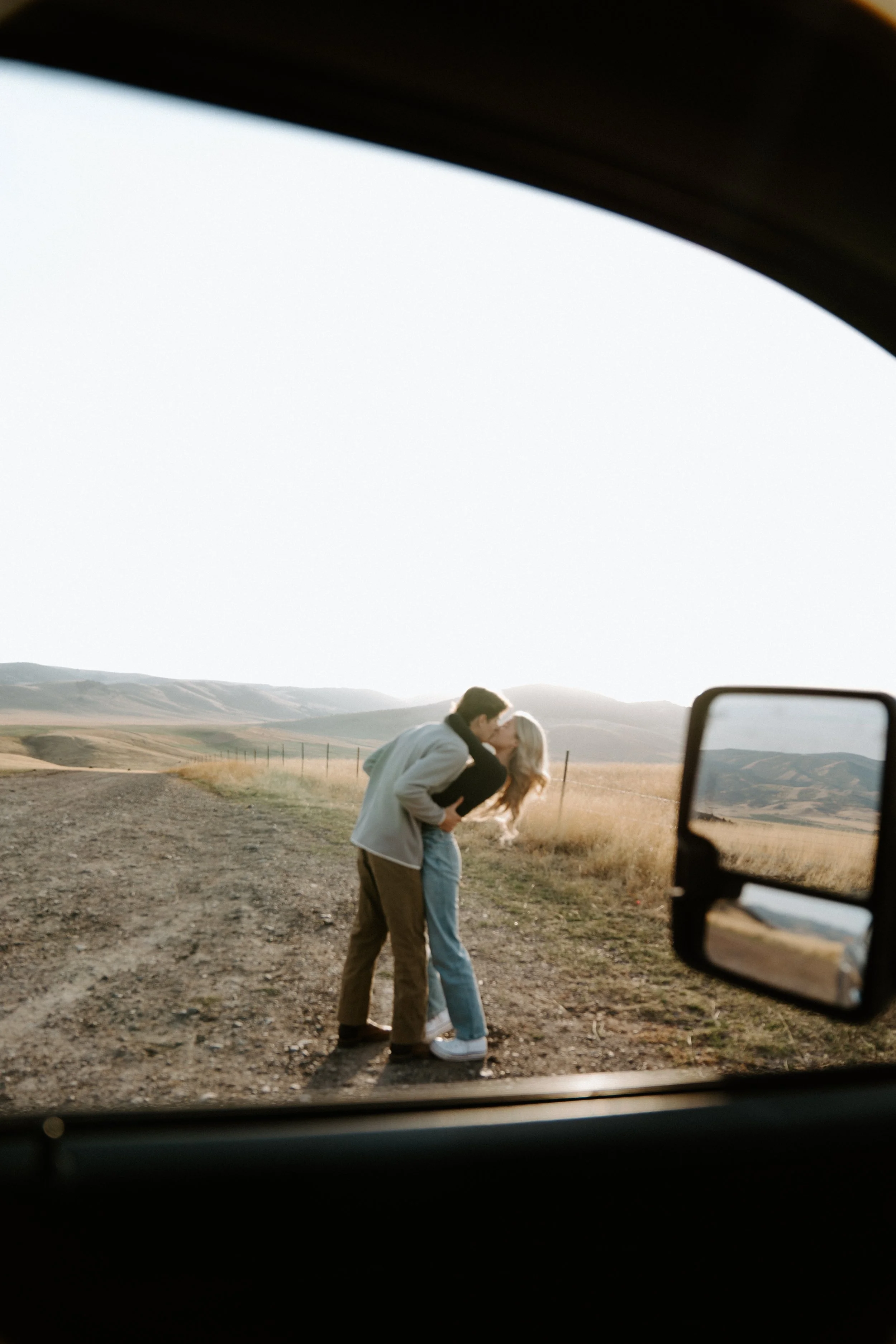 A couple sharing a kiss on a dirt road in a rural landscape, viewed through a car window with a side mirror, during sunset.