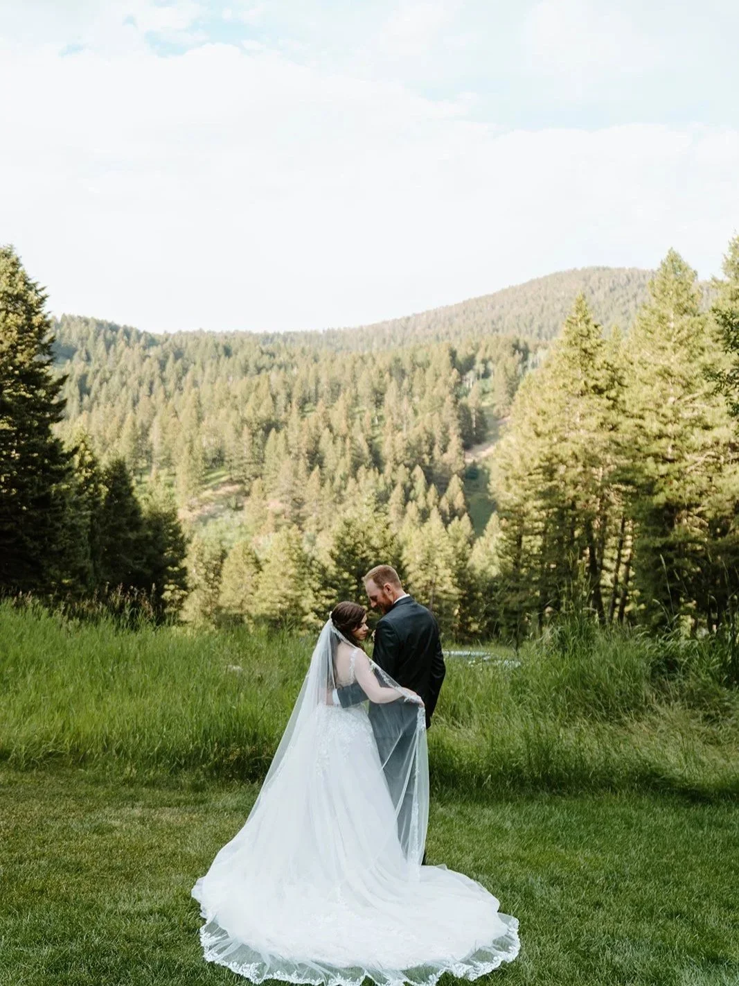 A bride and groom standing closely together on a grassy field in front of a forested mountain landscape.