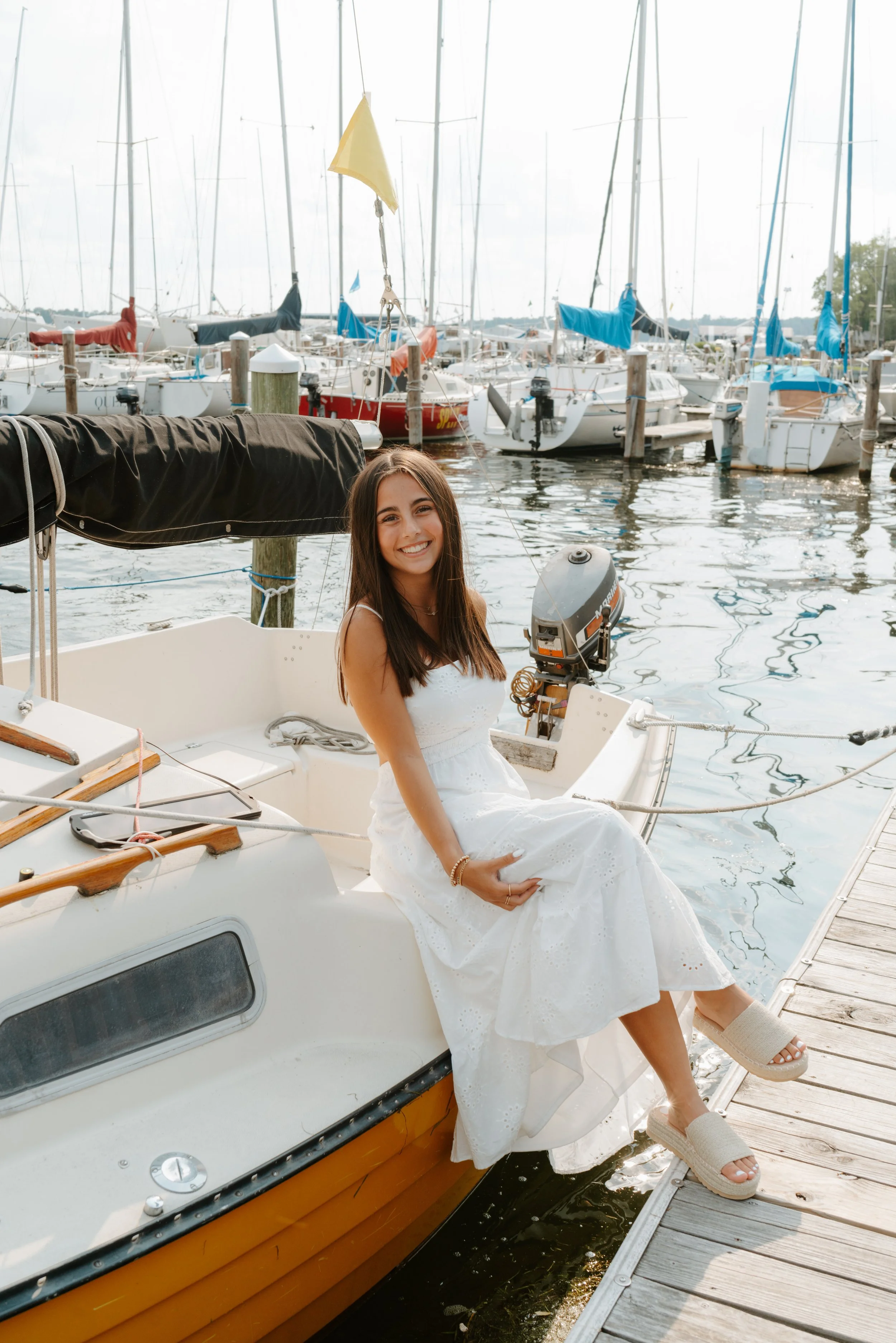 A young woman sitting on the edge of a white and yellow sailboat at a marina, smiling, wearing a white dress and sandals. Multiple sailboats are docked in the background.