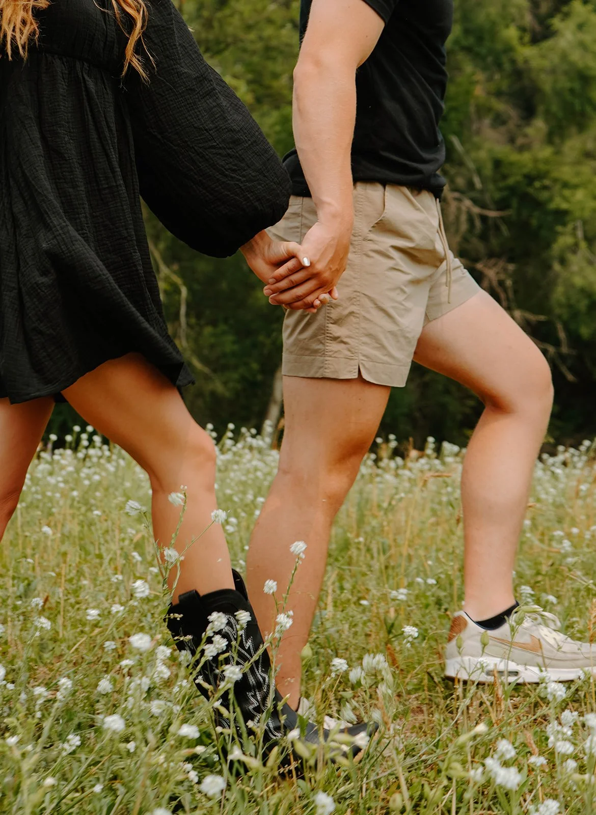 A couple walking hand in hand through a grassy field with small white flowers, holding hands, with trees in the background.