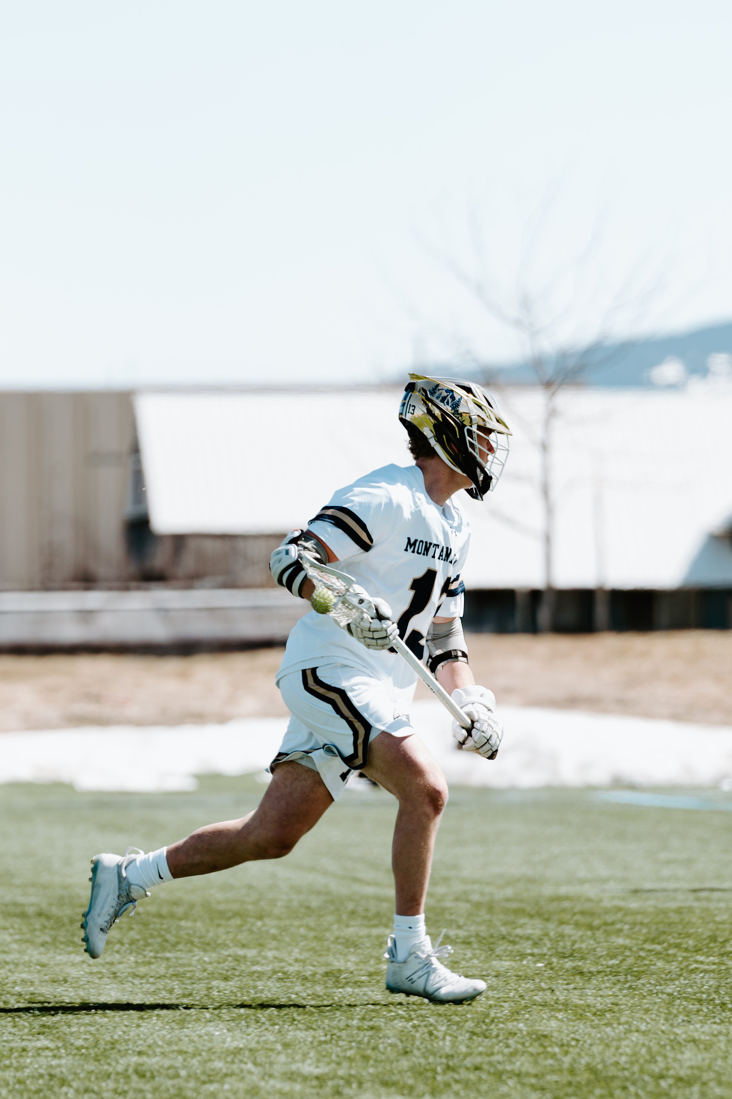 A person in a white hockey uniform running on a field while holding a lacrosse stick, wearing a helmet and gloves, with a background of buildings and trees.