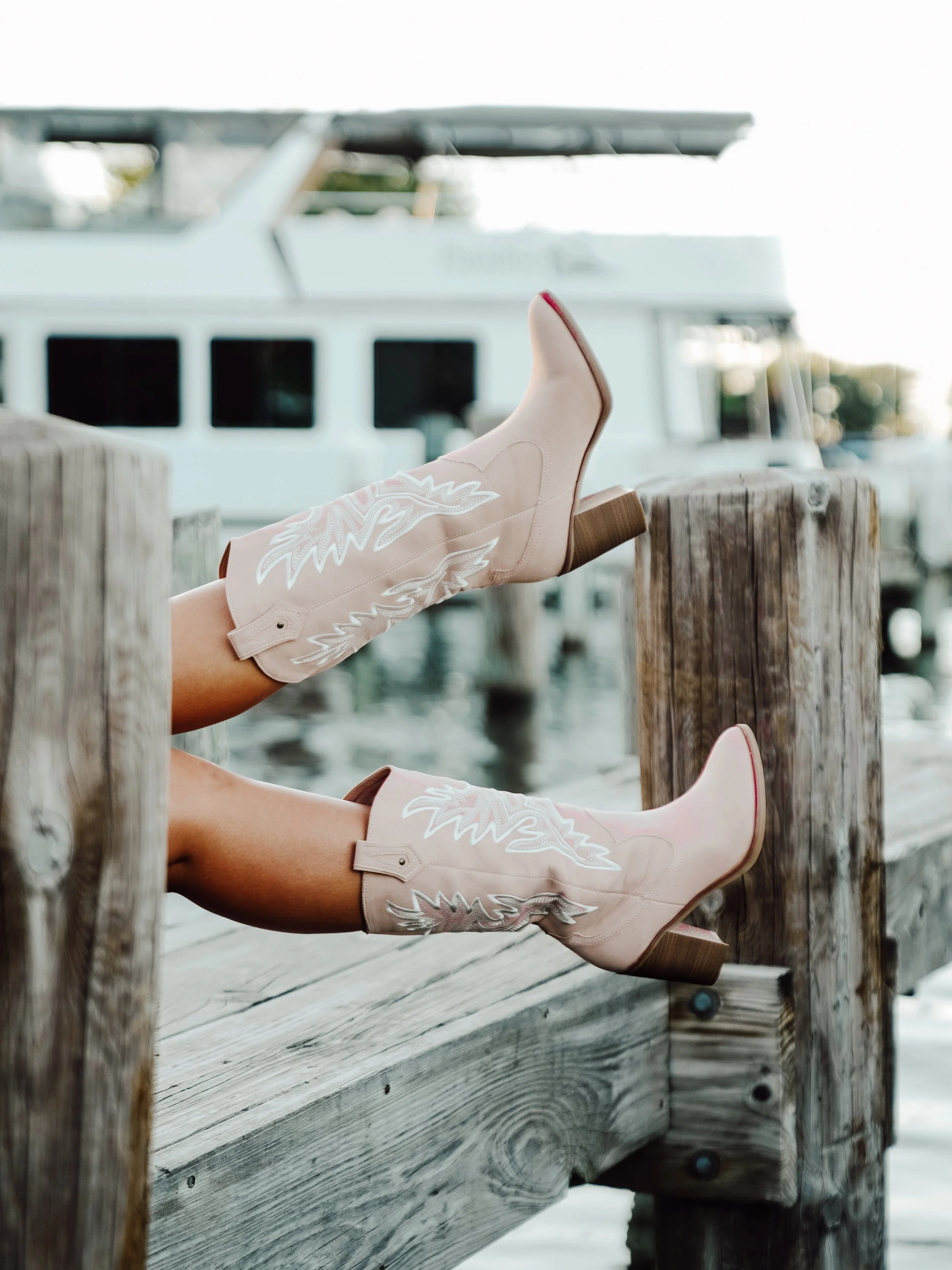 A person wearing light pink cowboy boots with white embroidered designs, lounging on a wooden dock near water, with boats in the background.