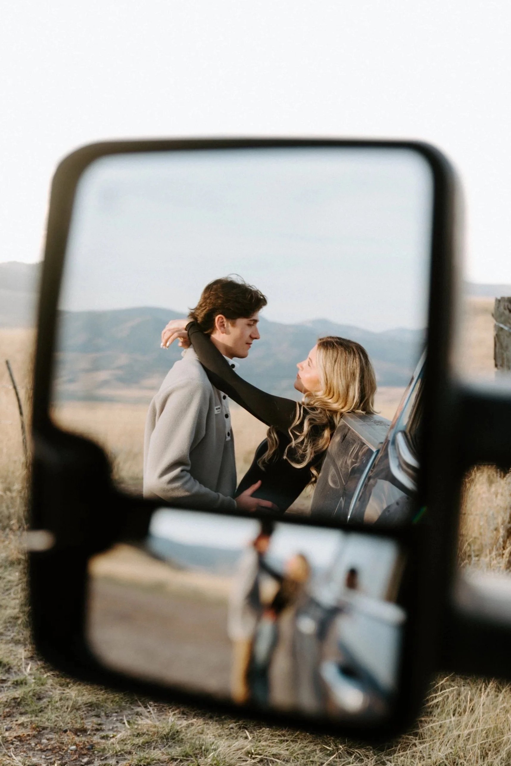 A man and a woman are visible through the side mirror of a vehicle, with the woman touching the man's face as they look at each other in a rural outdoor setting.