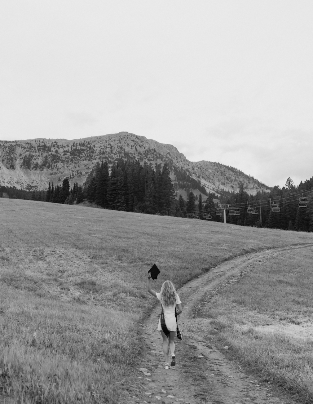 A woman walking on a dirt trail through grassy fields with a mountain and pine trees in the background, holding a jacket in her hand.