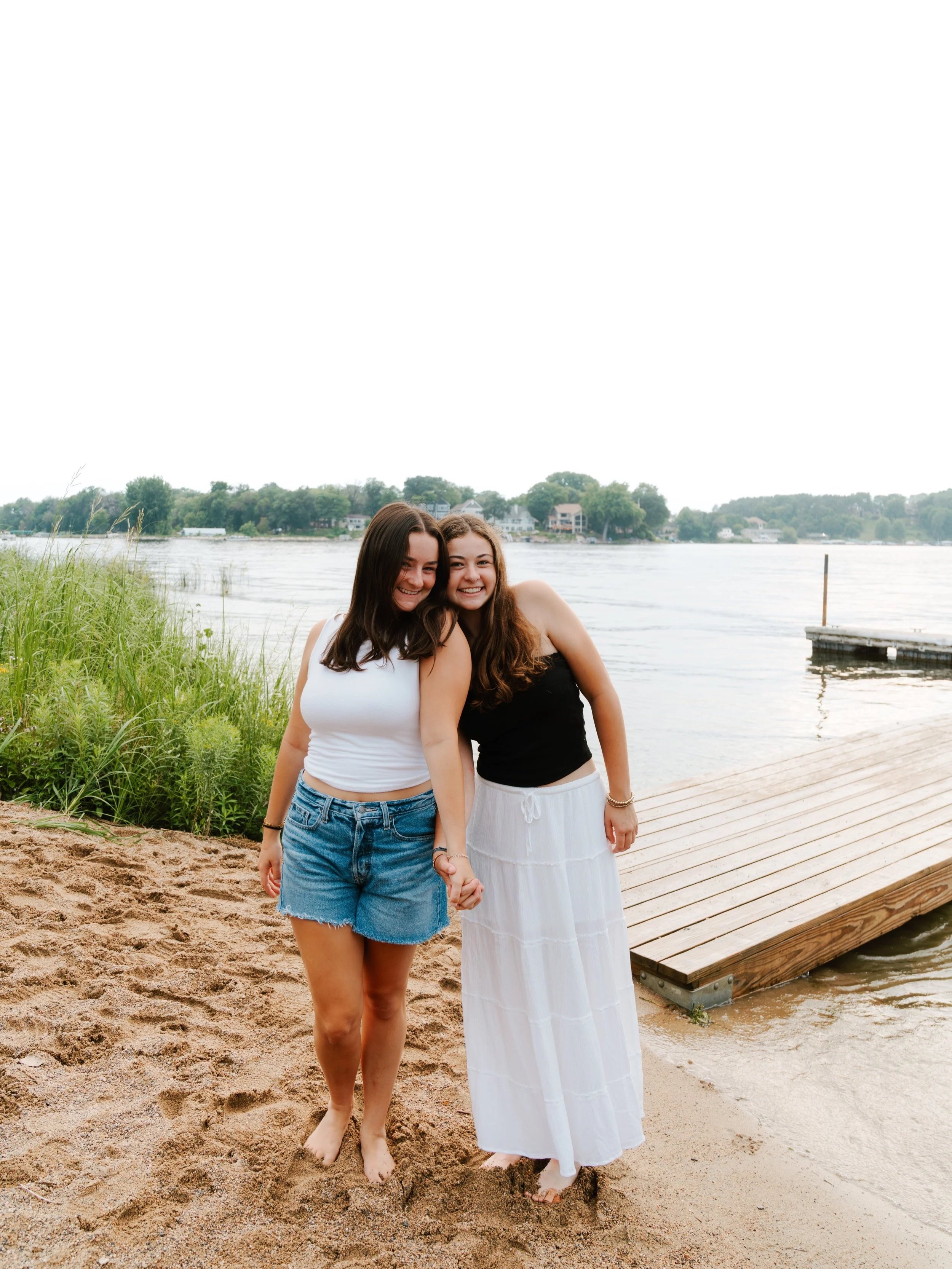 Two young women standing barefoot on a sandy beach near a wooden dock by a river, smiling and holding hands with greenery and houses in the background.