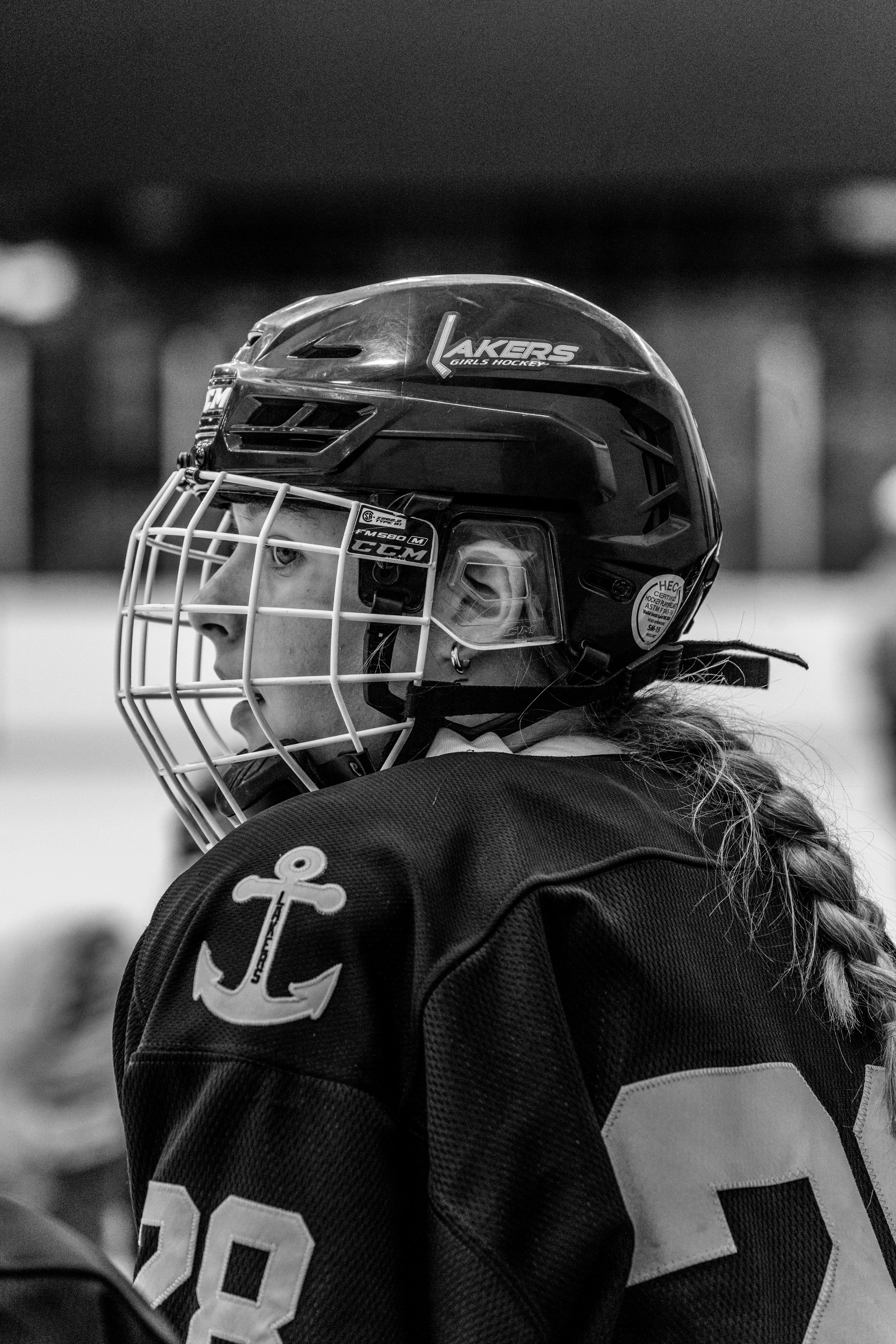 Black and white photo of a female ice hockey player wearing a helmet with a face cage and a jersey with the number 28 and an anchor emblem on the sleeve, looking to the left.