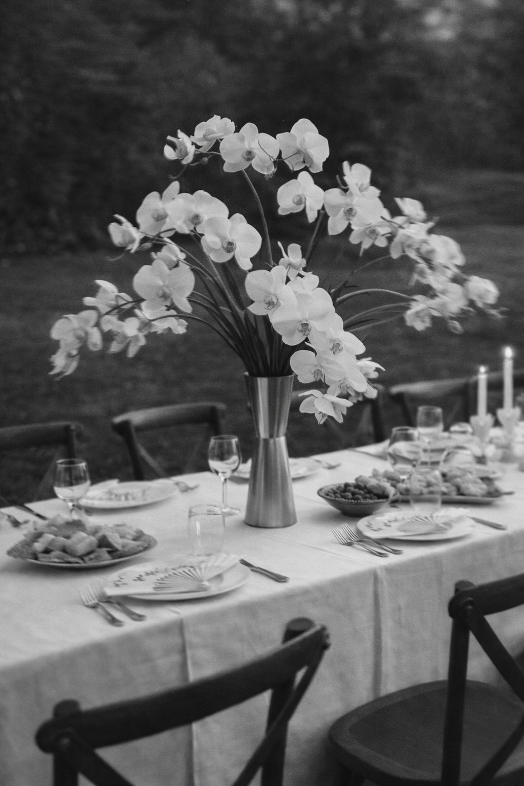 Ein festlich gedeckter Tisch mit einer großen Vase voller weißer Blumen, Tafelgeschirr, Gläsern und Kerzen im Hintergrund, wahrscheinlich bei einer Dinnerparty im Freien.