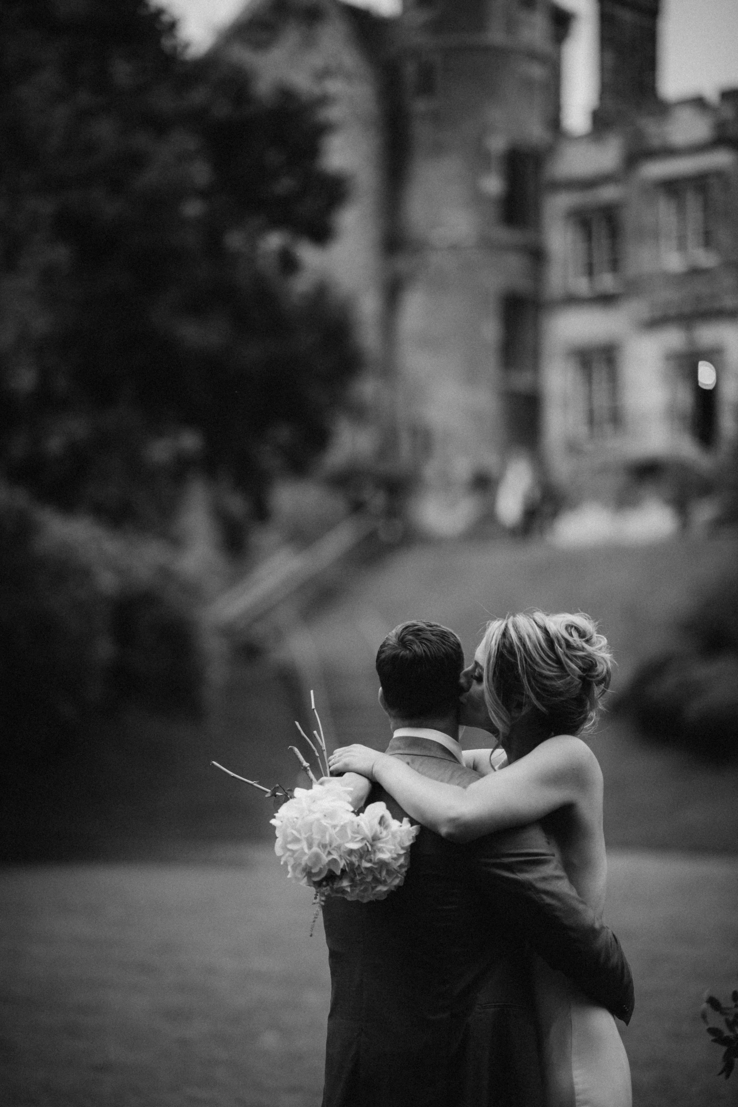 Bride and groom embracing outdoors, holding a bouquet, blurred background of a castle.