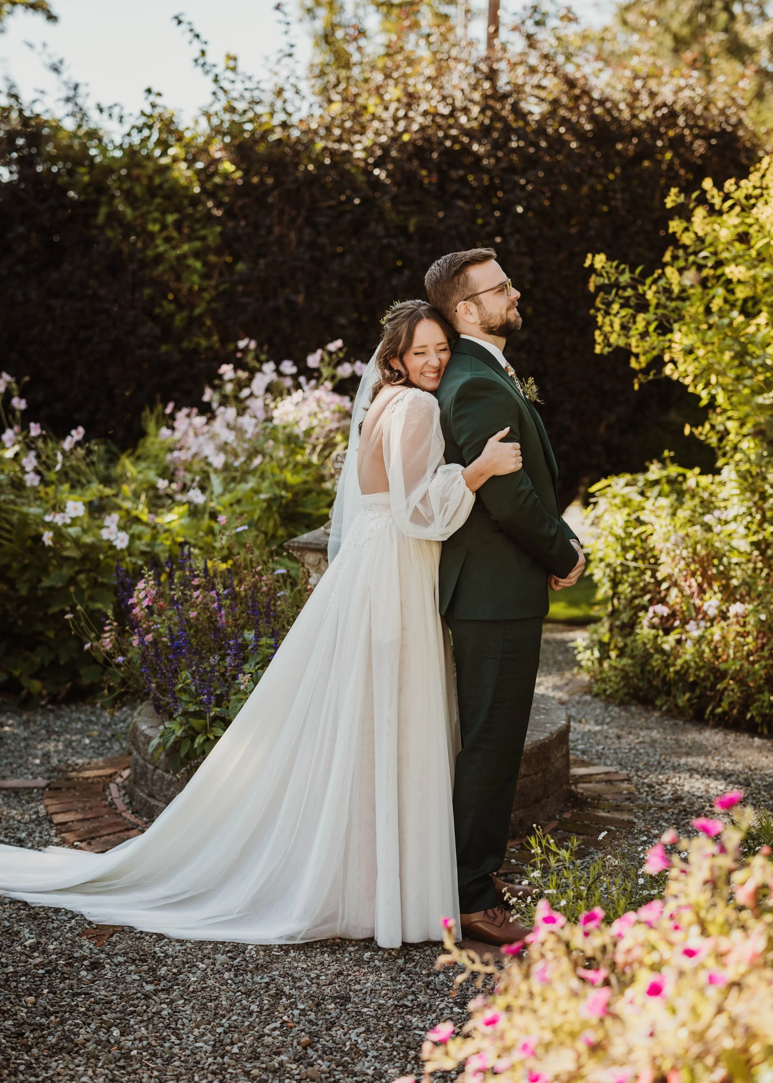 A bride in a flowing gown embraces her groom from behind during their garden first look. She smiles joyfully as he stands in anticipation, surrounded by lush greenery and blooming flowers. Captured by a Seattle wedding photographer at Snohomish venue