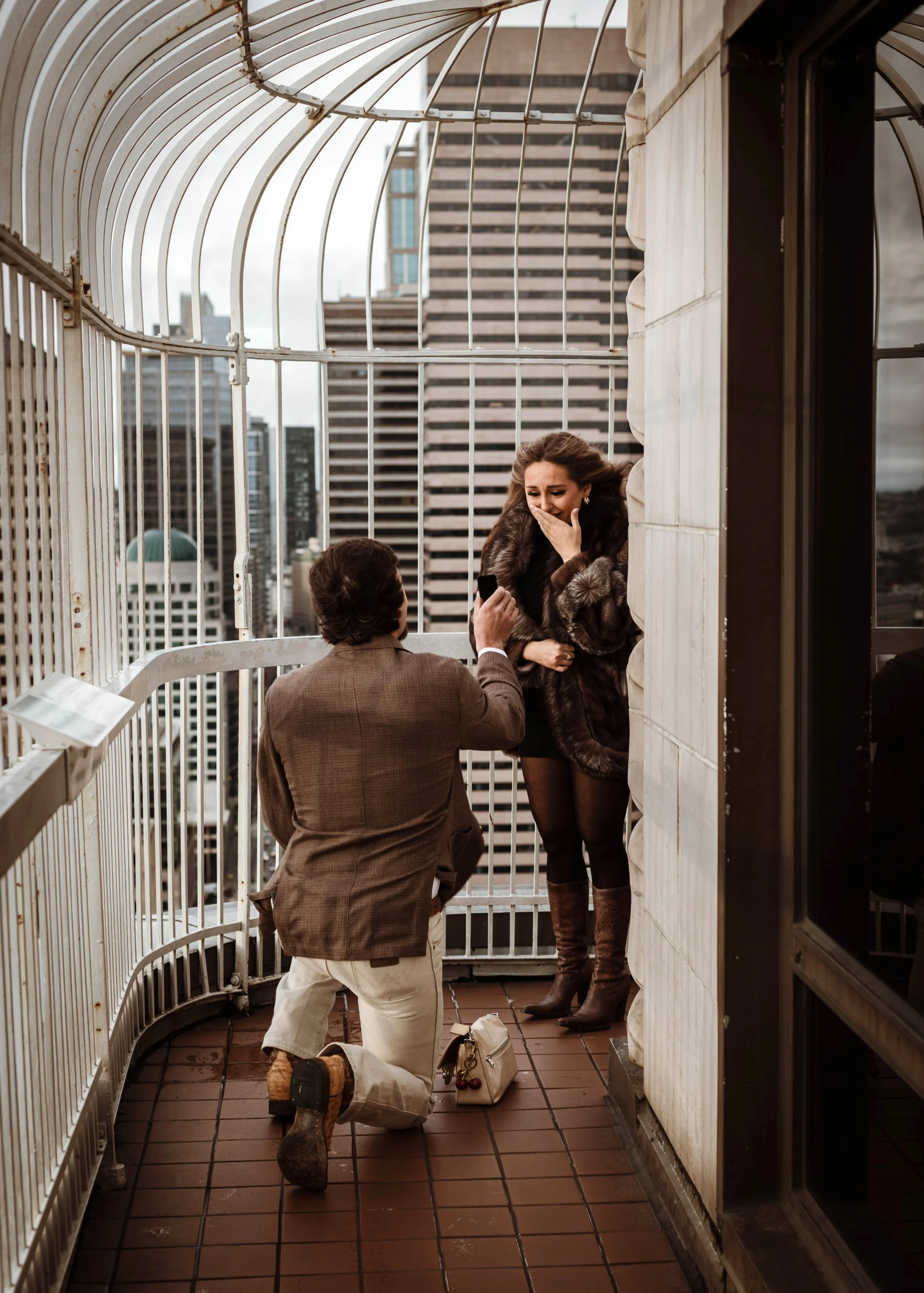Romantic rooftop proposal in downtown Seattle. A man kneels, holding out a ring as his partner, dressed in a fur coat and boots, covers her mouth in shock. City skyscrapers frame this unforgettable moment. Captured by a Seattle proposal photographer.