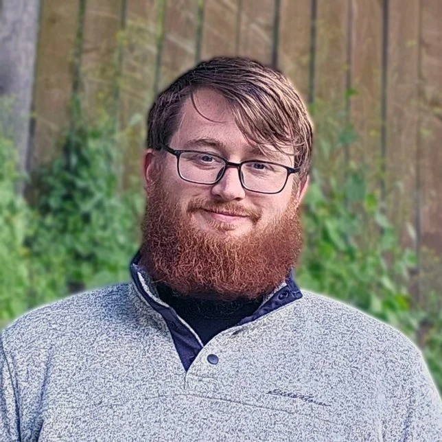 A man smiling outdoors in front of a wooden fence and green foliage.