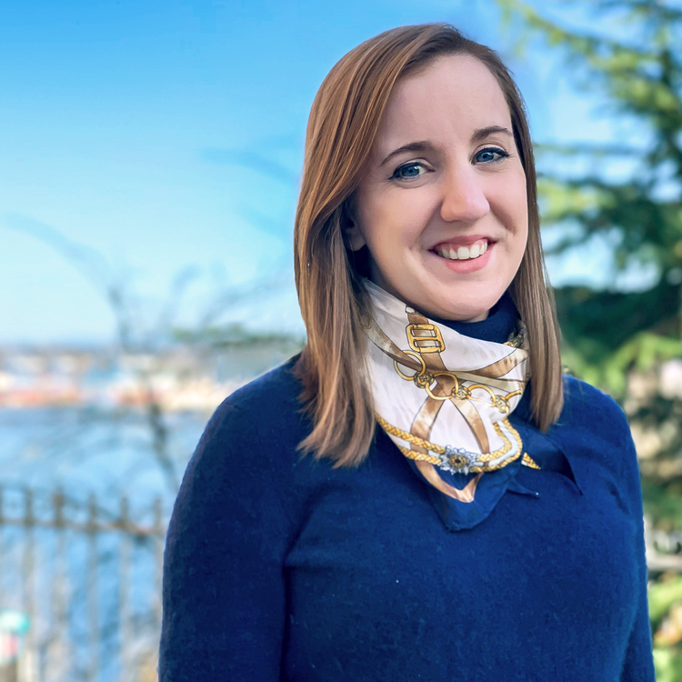A woman with shoulder-length brown hair smiling outdoors, wearing a navy blue coat and a patterned scarf, with a blurred background of trees and water.