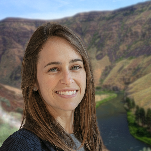 A woman smiling outdoors with a landscape of hills, a river, and a partly cloudy sky.