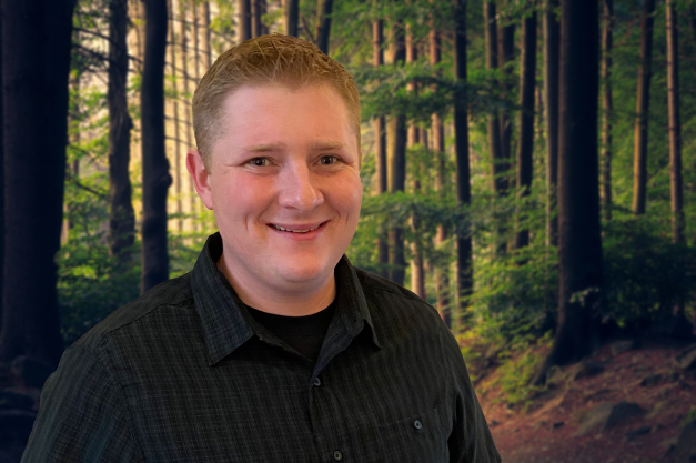 A smiling man with short blonde hair wearing a black shirt stands in front of a forest.