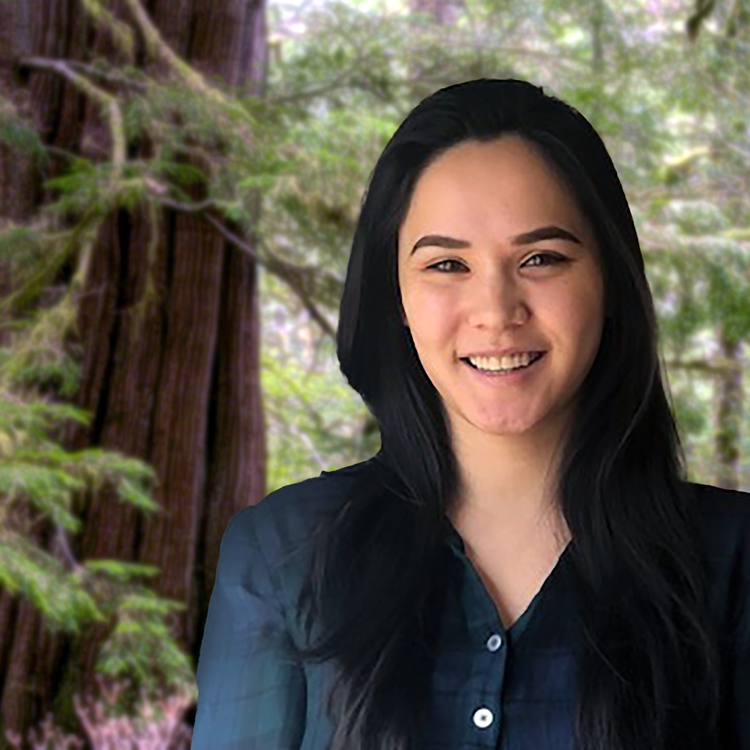 A woman with long black hair smiling, wearing a dark shirt, standing outdoors in a forest.