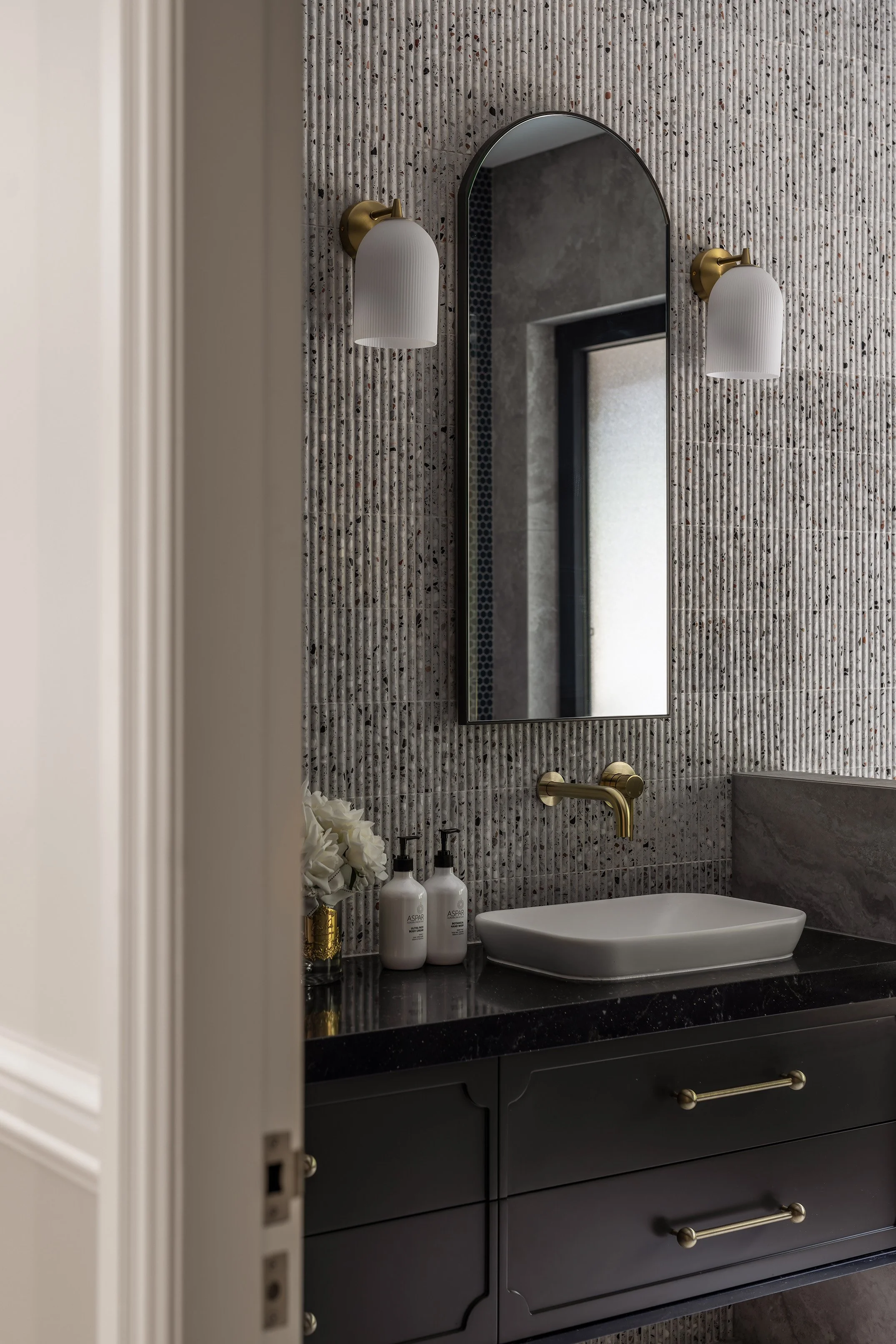 A modern bathroom vanity with a black countertop, a white rectangular vessel sink, a gold faucet, and two wall sconces with white shades. A mirror is mounted on a textured, patterned wall, with a window reflected in it. Two soap dispensers and a smal