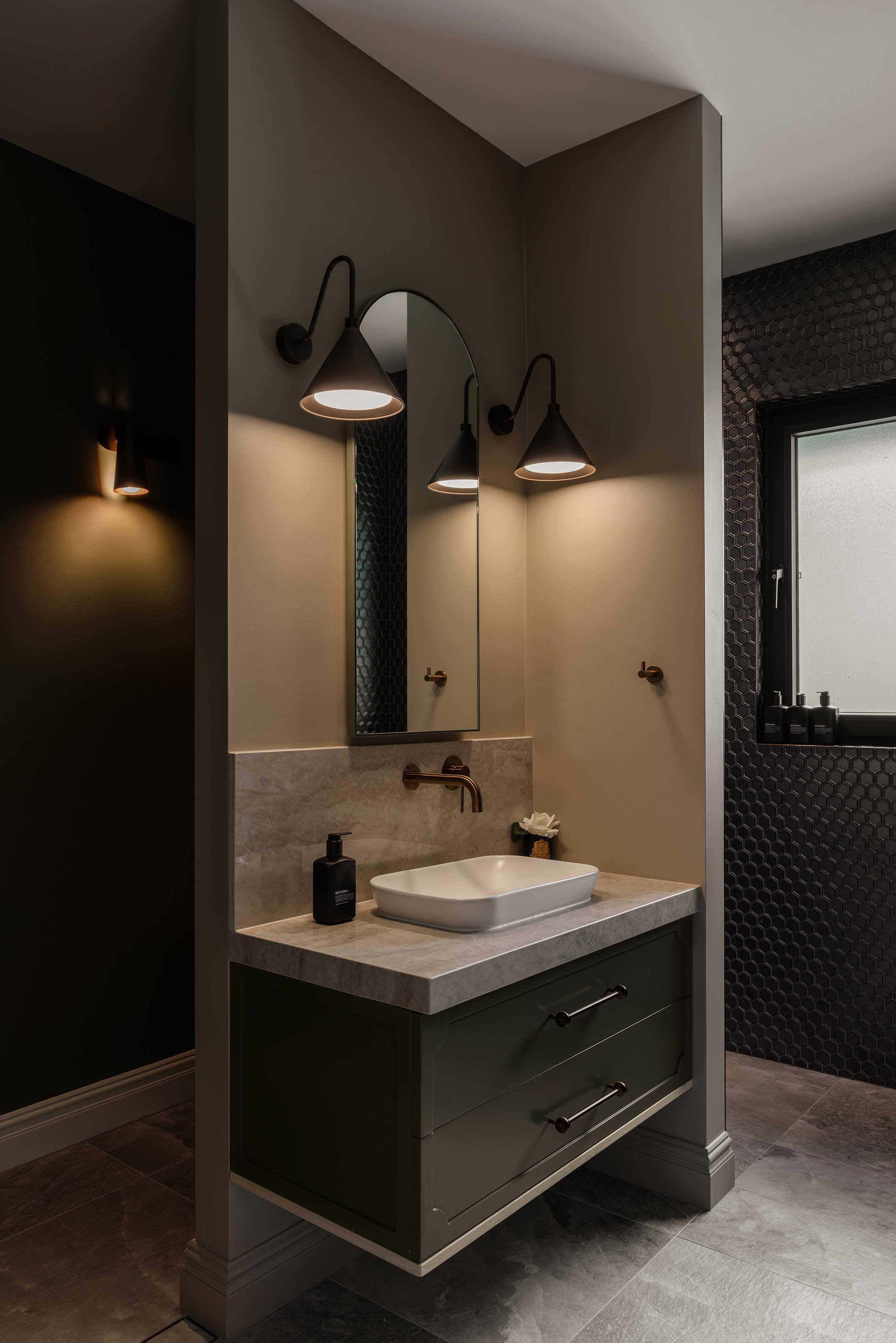 Modern bathroom with a green vanity, marble countertop, vessel sink, wall-mounted faucet, oval mirror, black wall sconces, and a frosted window with black trim.