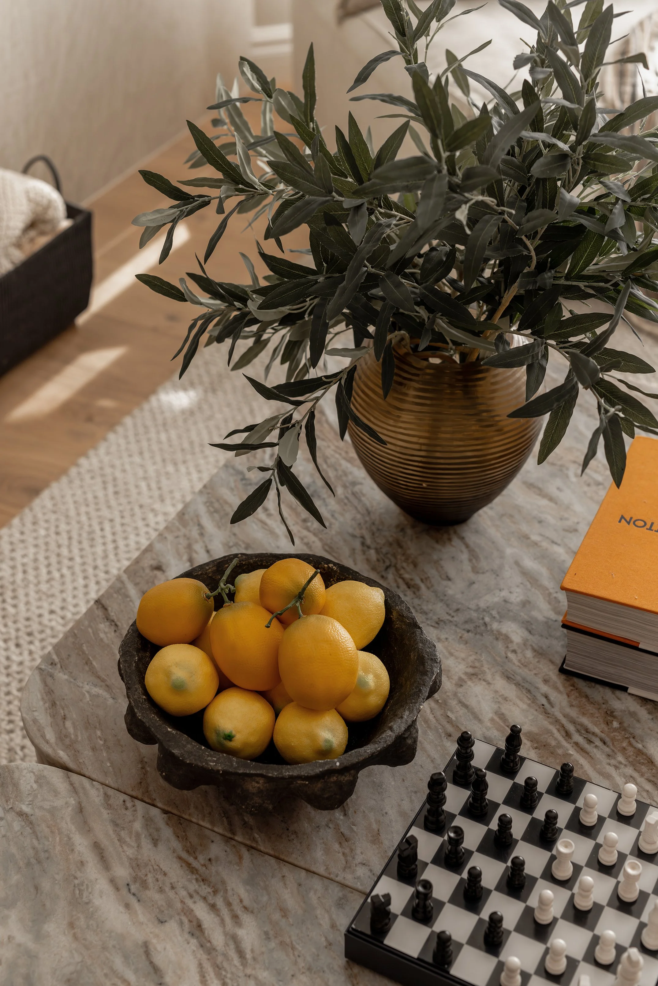 A marble table with a bowl of lemons, a potted plant with green leaves in a striped brown vase, books, and a chessboard.