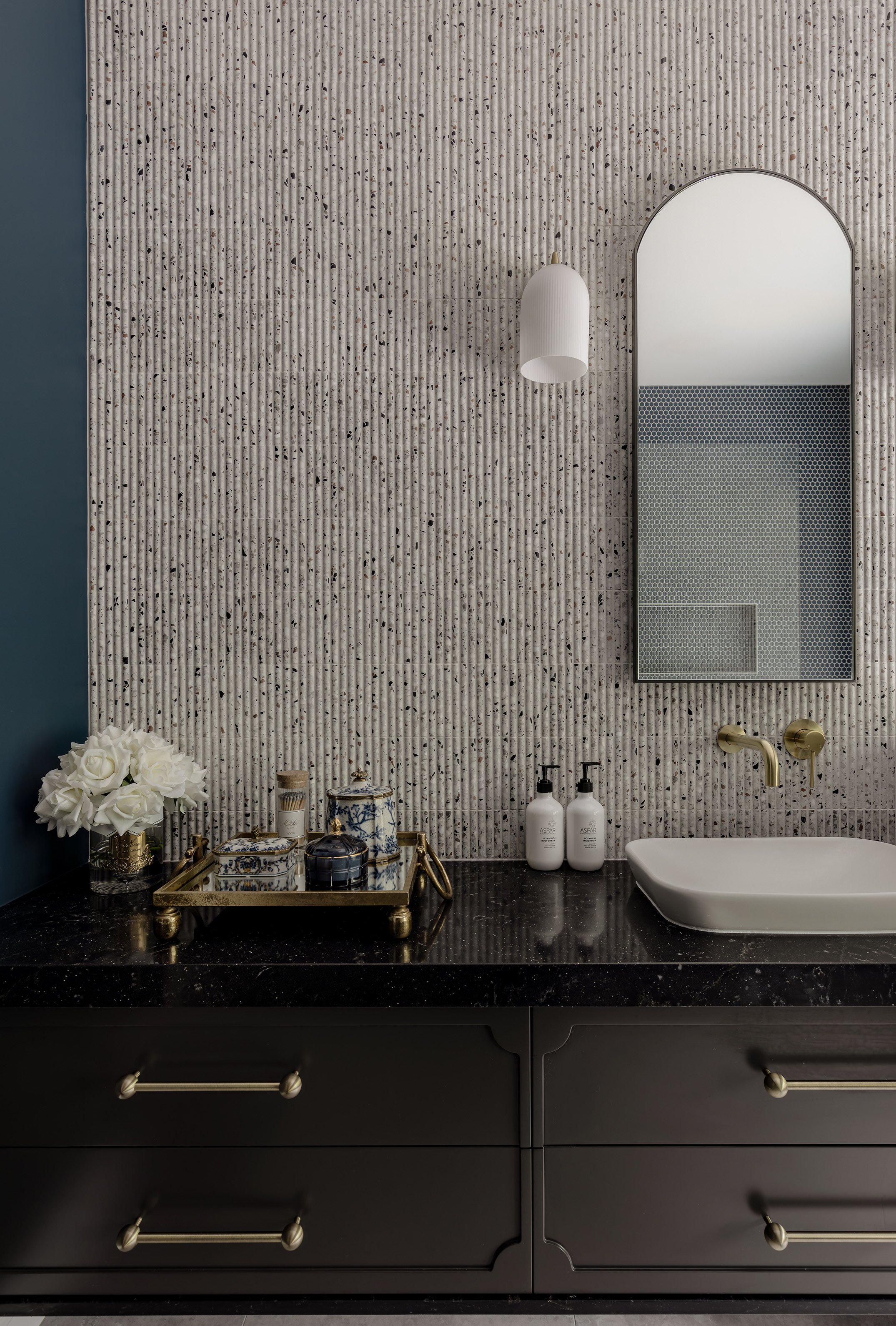 Modern bathroom vanity with a black countertop, white sink, brass fixtures, a mirror, white soap dispensers, and decorative items including a vase of white roses, a gold tray, and jars.