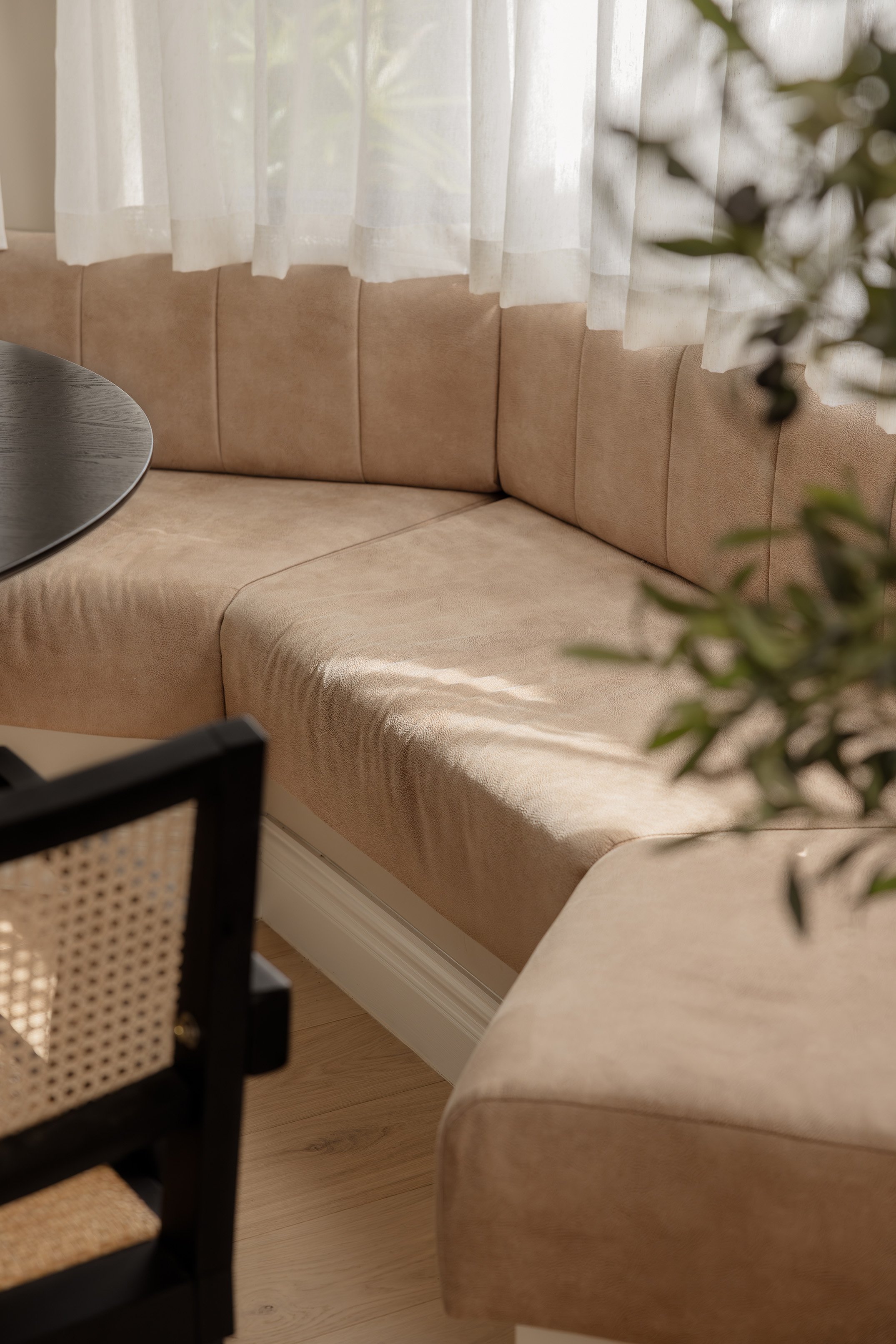 Beige upholstered corner bench seat by a window with sheer white curtains, next to a black table and a black chair with a woven rattan backrest in a cozy dining area.