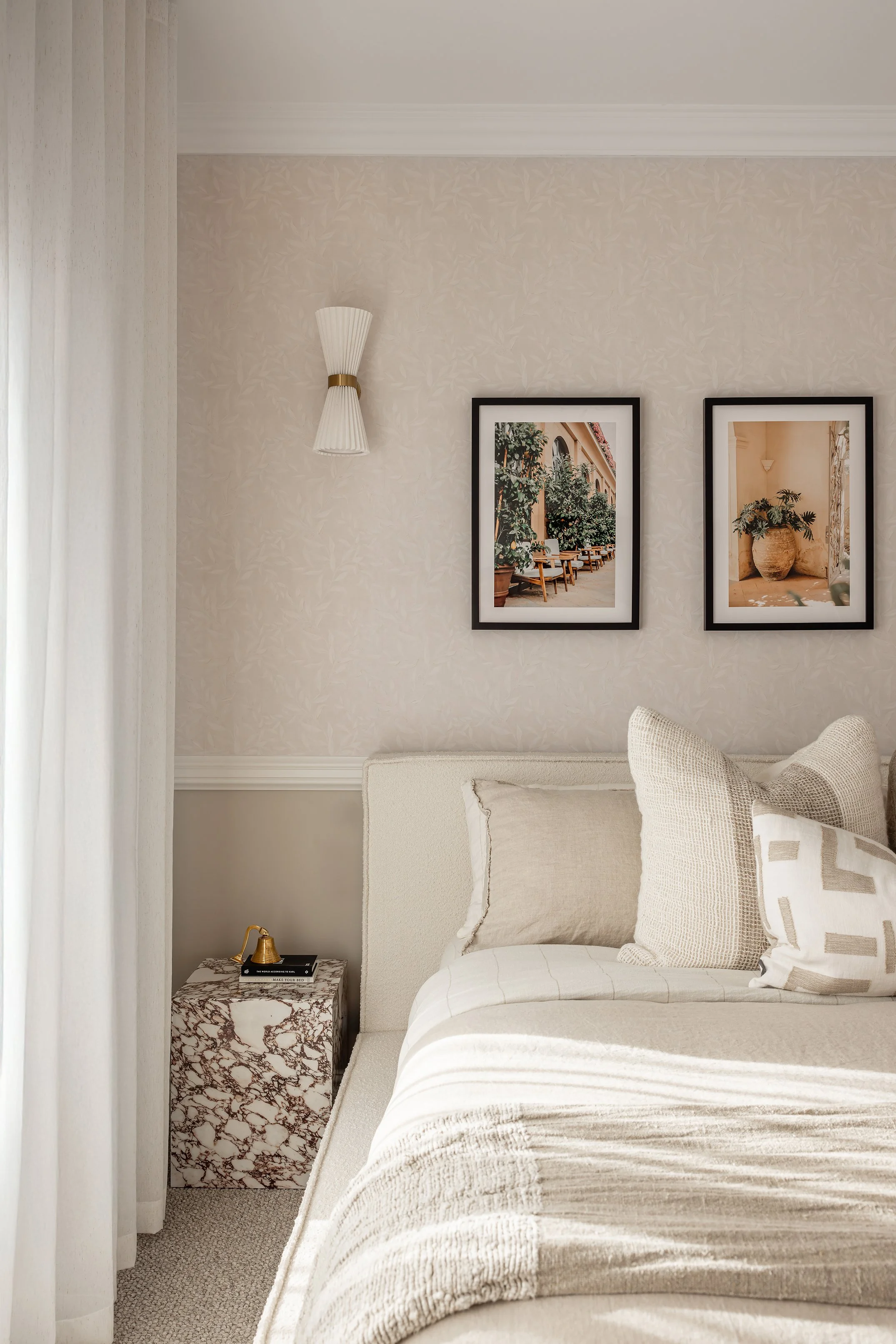 A cozy bedroom corner with beige and cream decor, including a bed with beige pillows and a textured blanket, a marble nightstand with a small brass lamp, and two framed artworks on the wall depicting outdoor scenes.