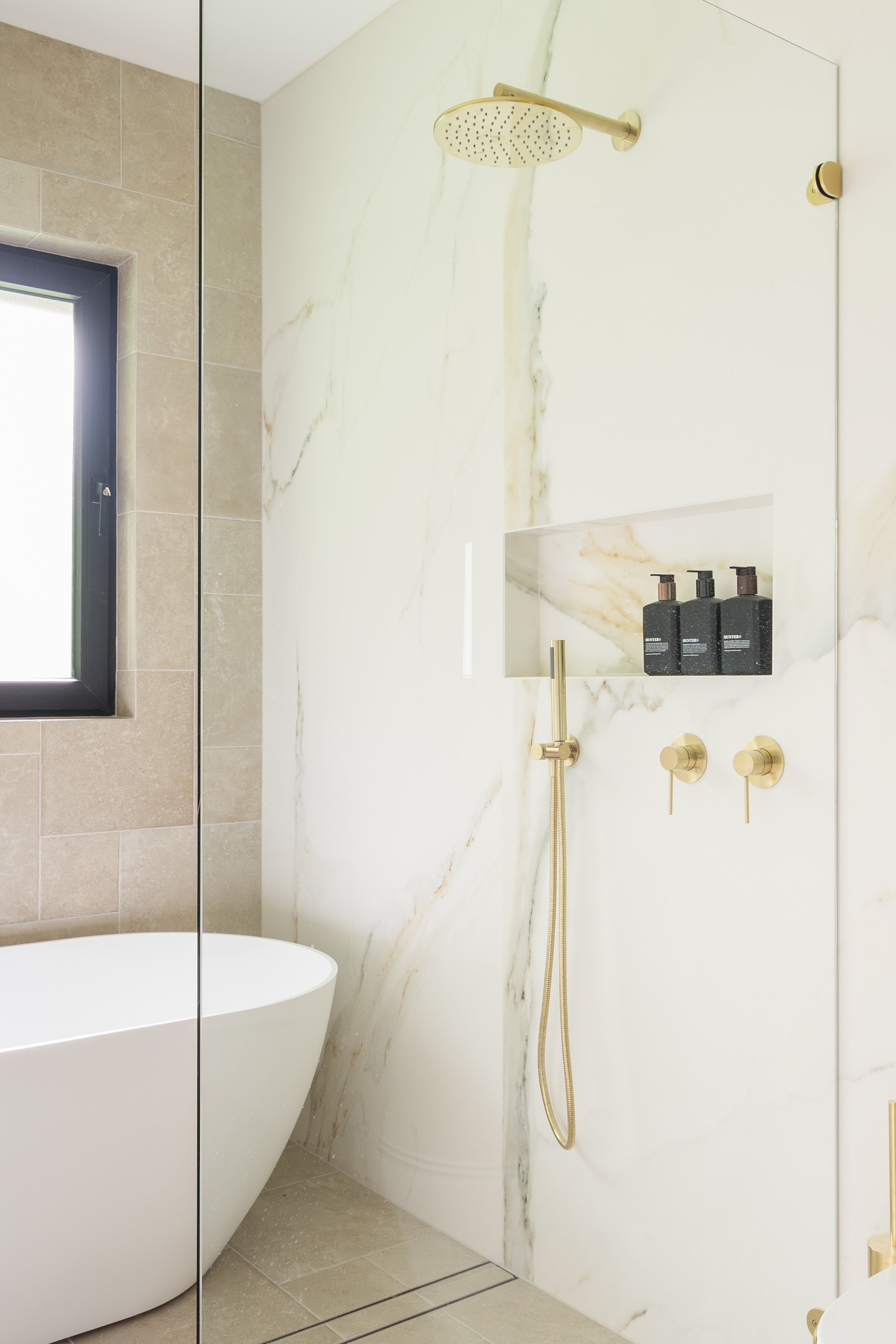 Modern bathroom with a white bathtub, beige tile walls, a black-framed window, and a marble shower wall with gold fixtures, including a rain showerhead and a handheld shower wand, with black soap dispensers in a built-in niche.