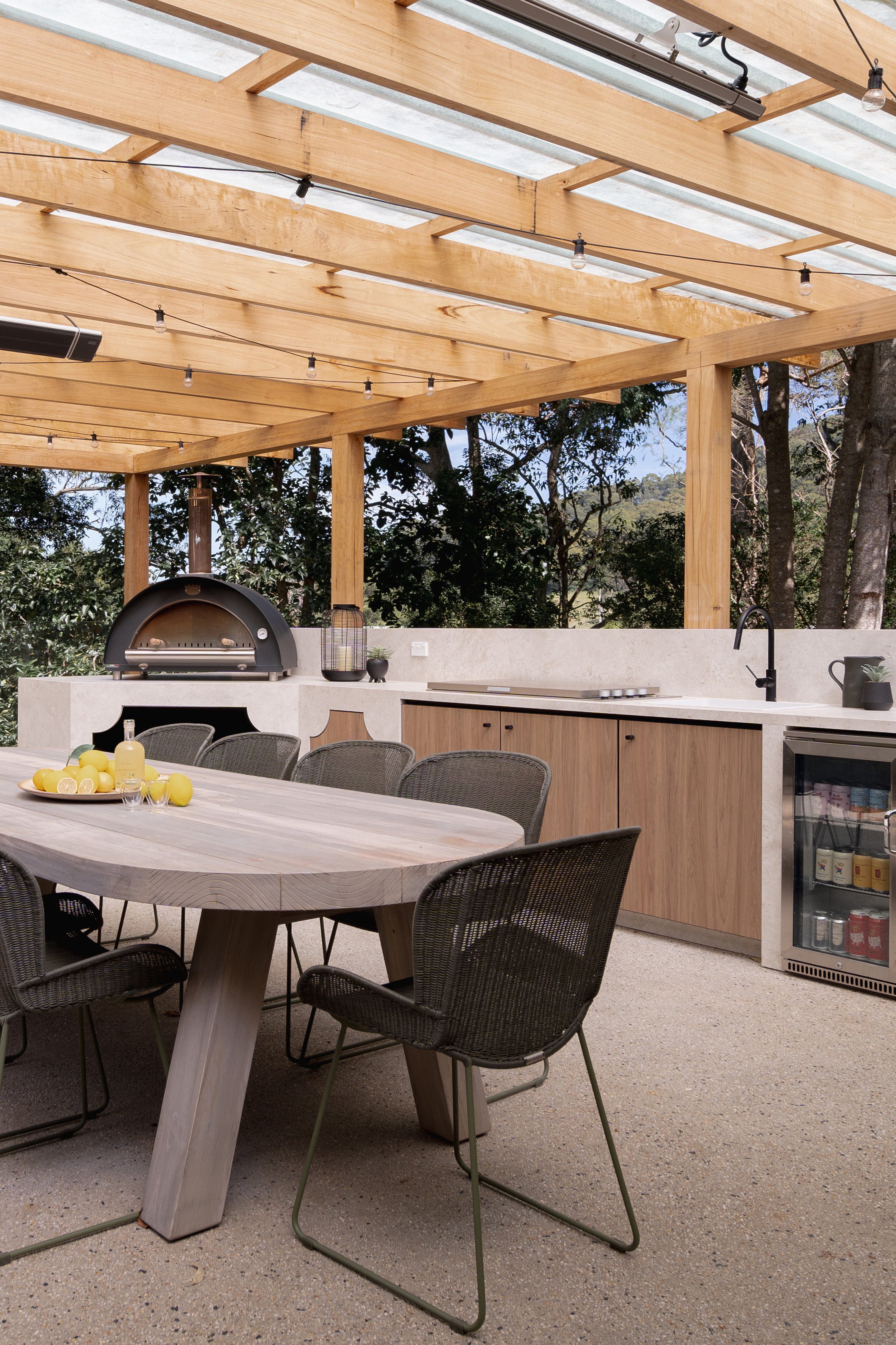 Outdoor covered patio area with a wooden ceiling, string lights, a dining table with black chairs, an outdoor pizza oven, a small refrigerator, and a black faucet outside.