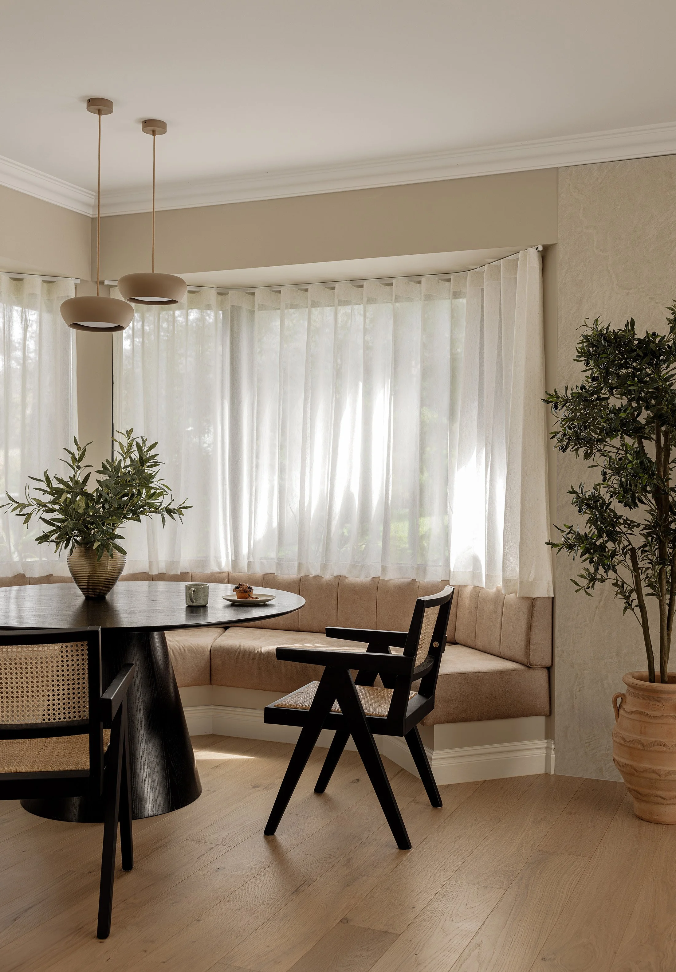 A cozy dining nook with a curved beige banquette, a dark wood round table with a large vase of green foliage, black chairs, sheer white curtains, and two modern pendant lights hanging from the ceiling.