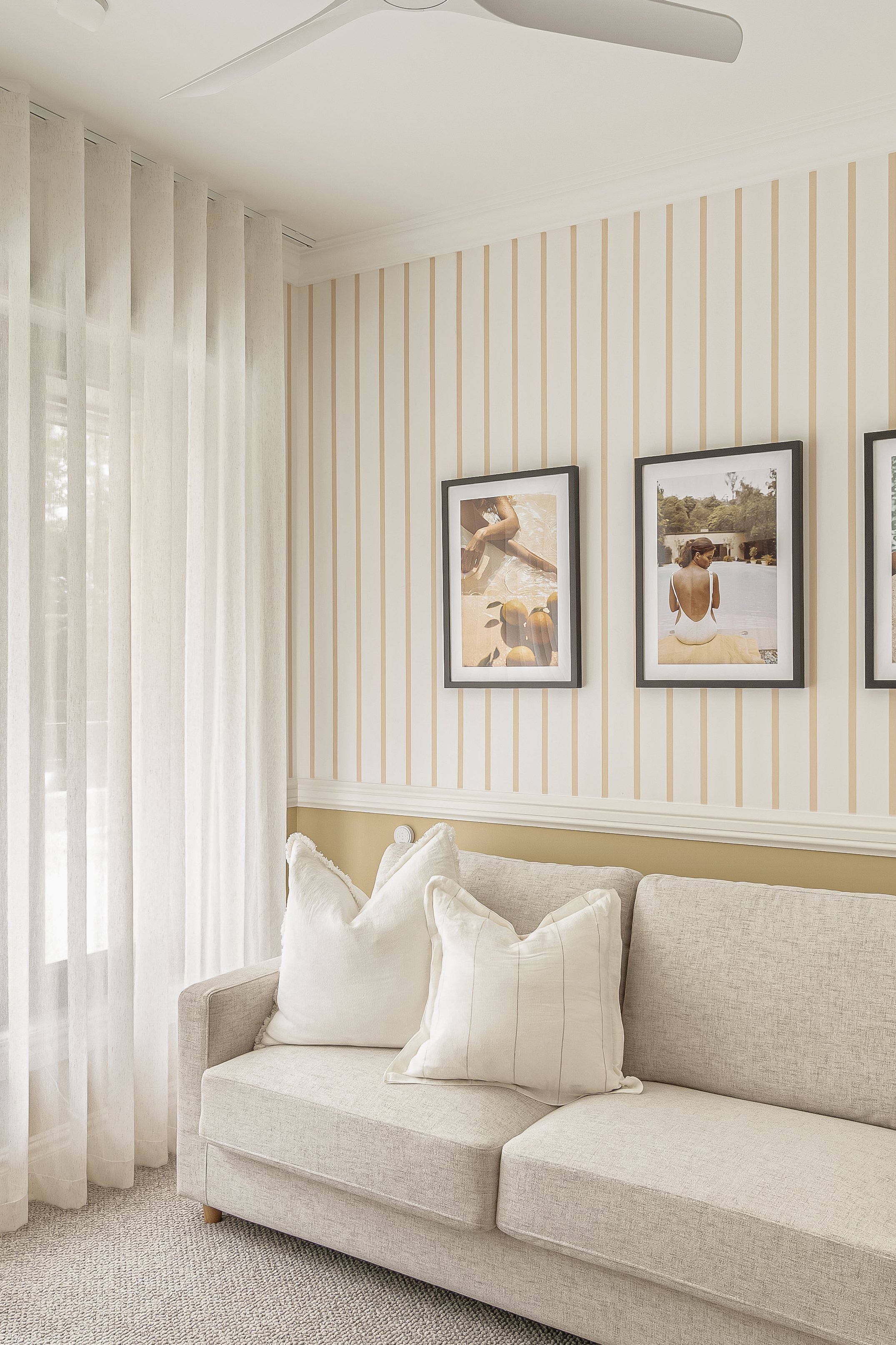 A living room corner with a beige sofa and decorative pillows, beige/sand striped wallpaper, framed photos on the wall, and sheer curtains letting in natural light.