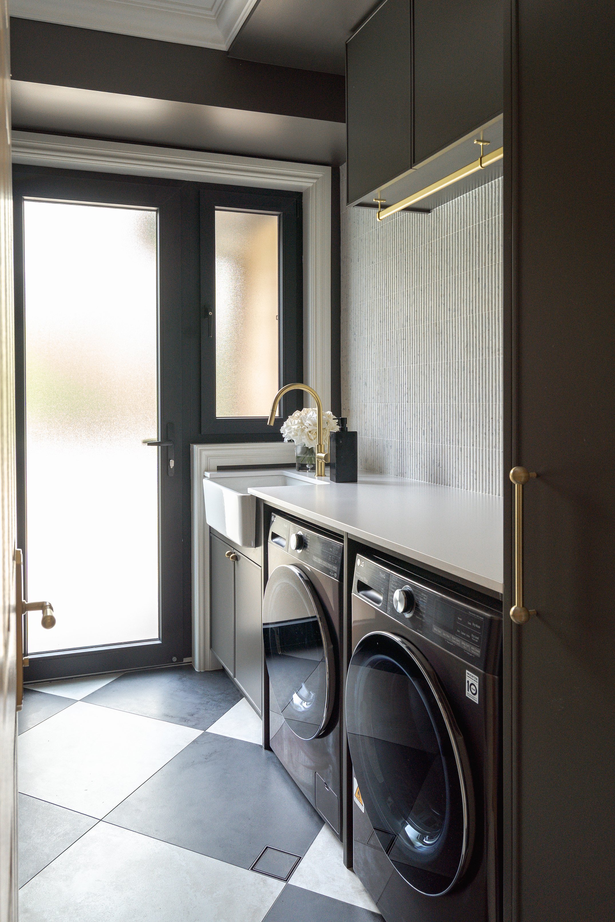 A modern laundry room with black and white checkered floor tiles, a white countertop, a gold faucet, a white laundry sink, two black front-loading laundry machines, a frosted glass door, a window, and dark upper cabinets.