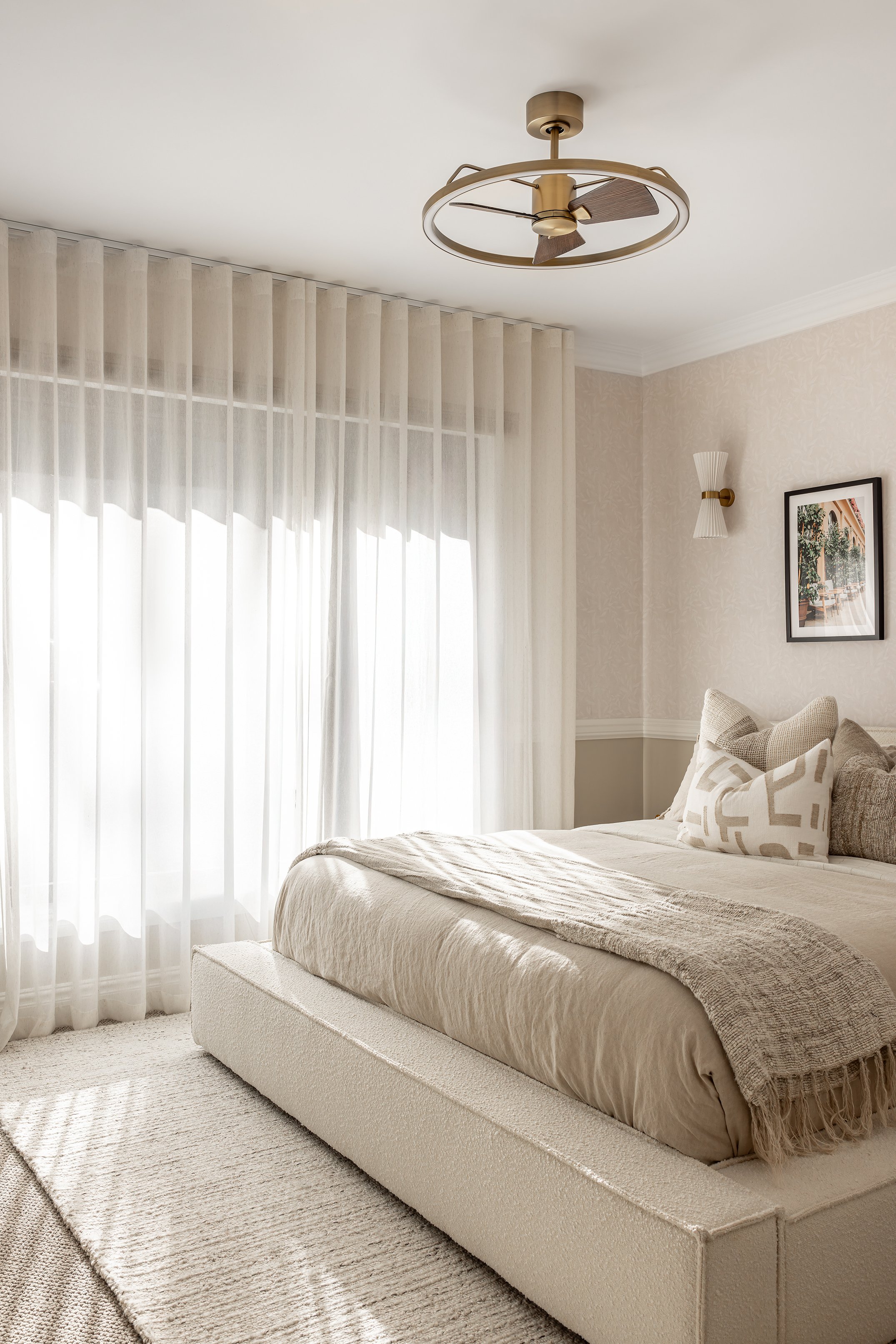 A bedroom with a beige bed and a large window with sheer white curtains, a ceiling fan, and wall-mounted light fixtures.