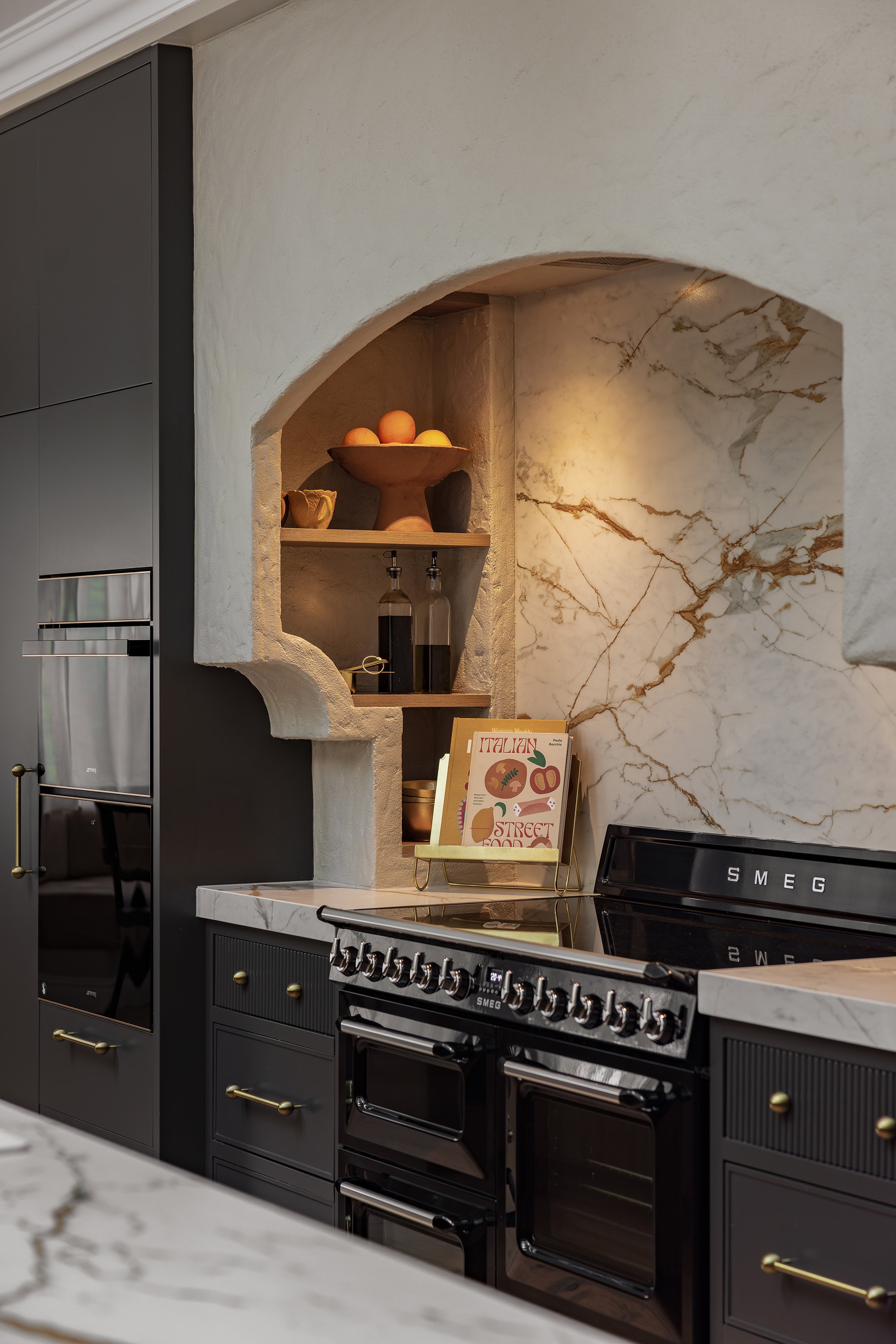 Kitchen with dark cabinetry, marble countertops, and a built-in marble arch with decorative shelves holding vases, books, and bottles. Features a black SMEG dual oven.