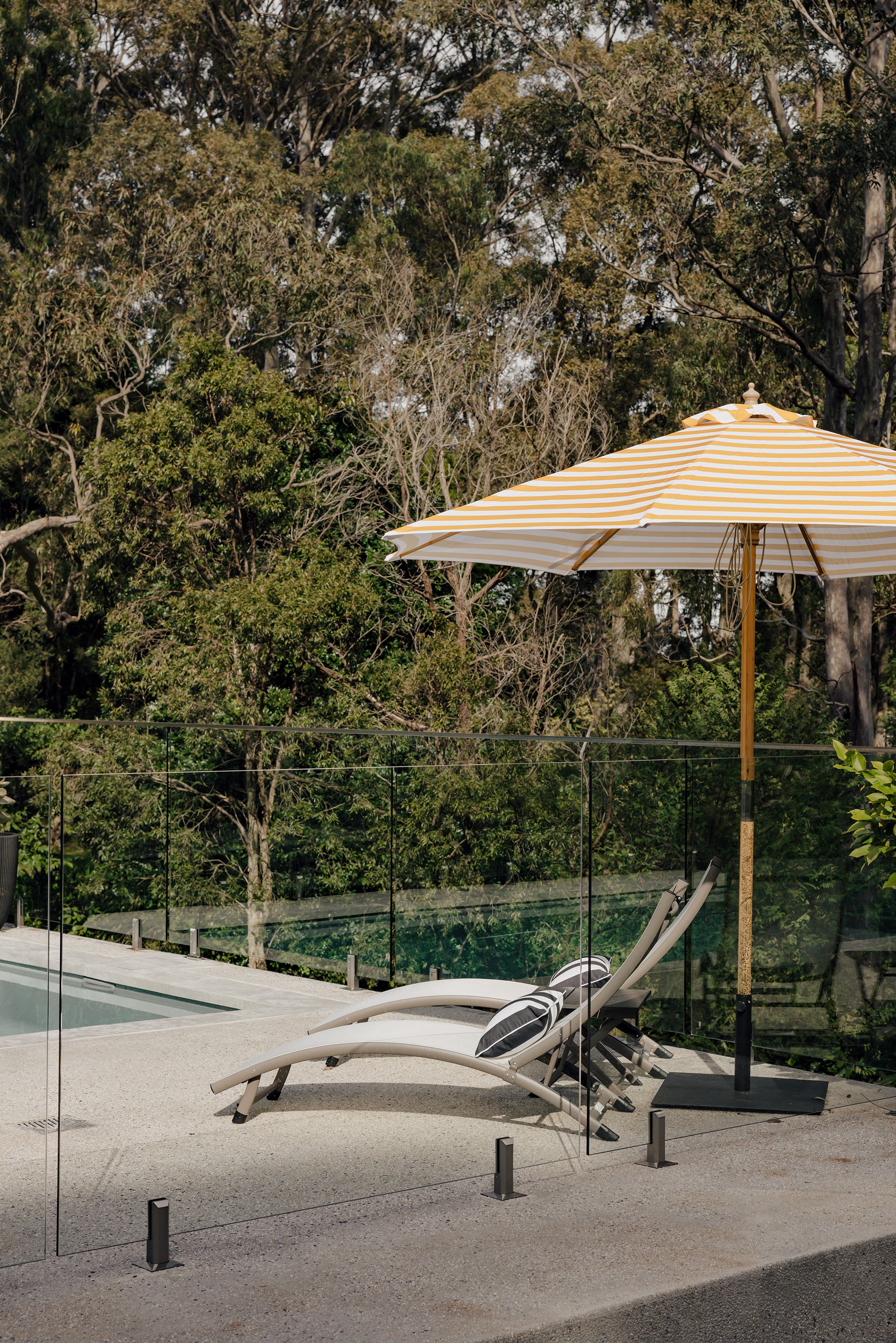 Poolside scene with two lounge chairs, black-striped pillows, a yellow and white striped umbrella, glass railing, and trees in the background.