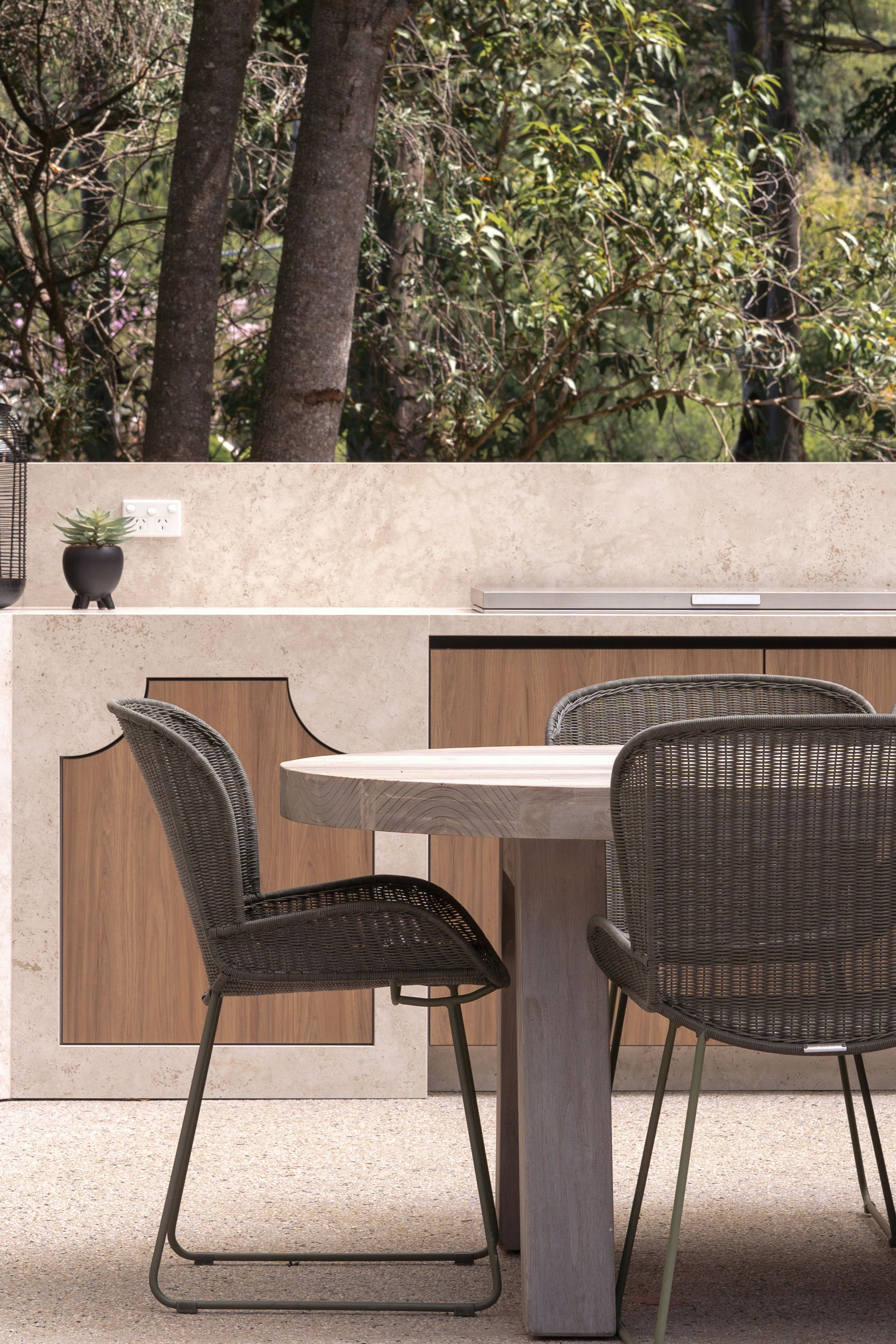 Modern indoor dining area with a round wooden table, two black wicker chairs, and a countertop with a small potted succulent, overlooking a garden view through a large window.