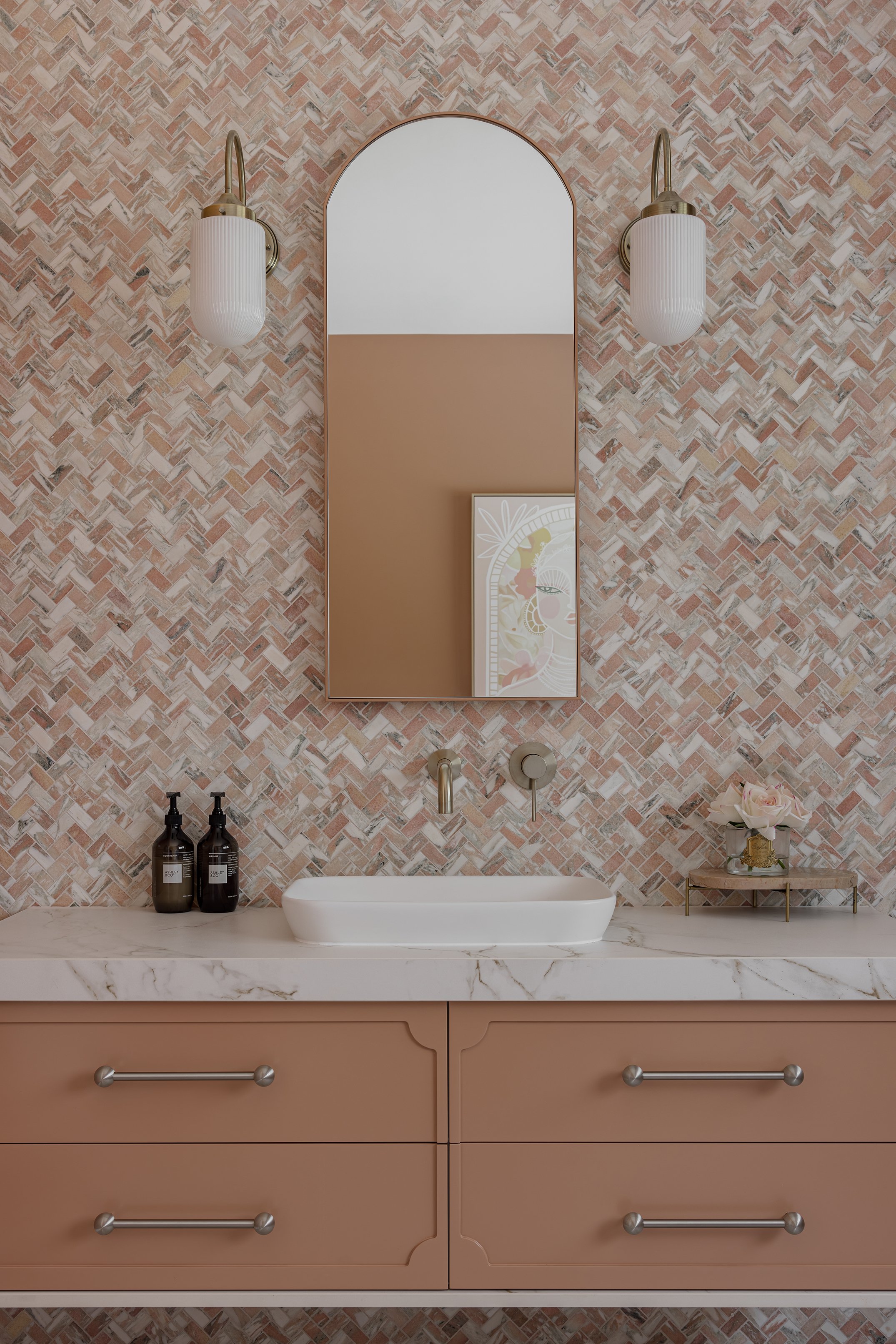 Bathroom vanity with marble countertop, white vessel sink, beige cabinet drawers, decorative wall mirror, wall-mounted faucets, two wall-mounted light fixtures, black soap dispensers, and a small tray with flowers.