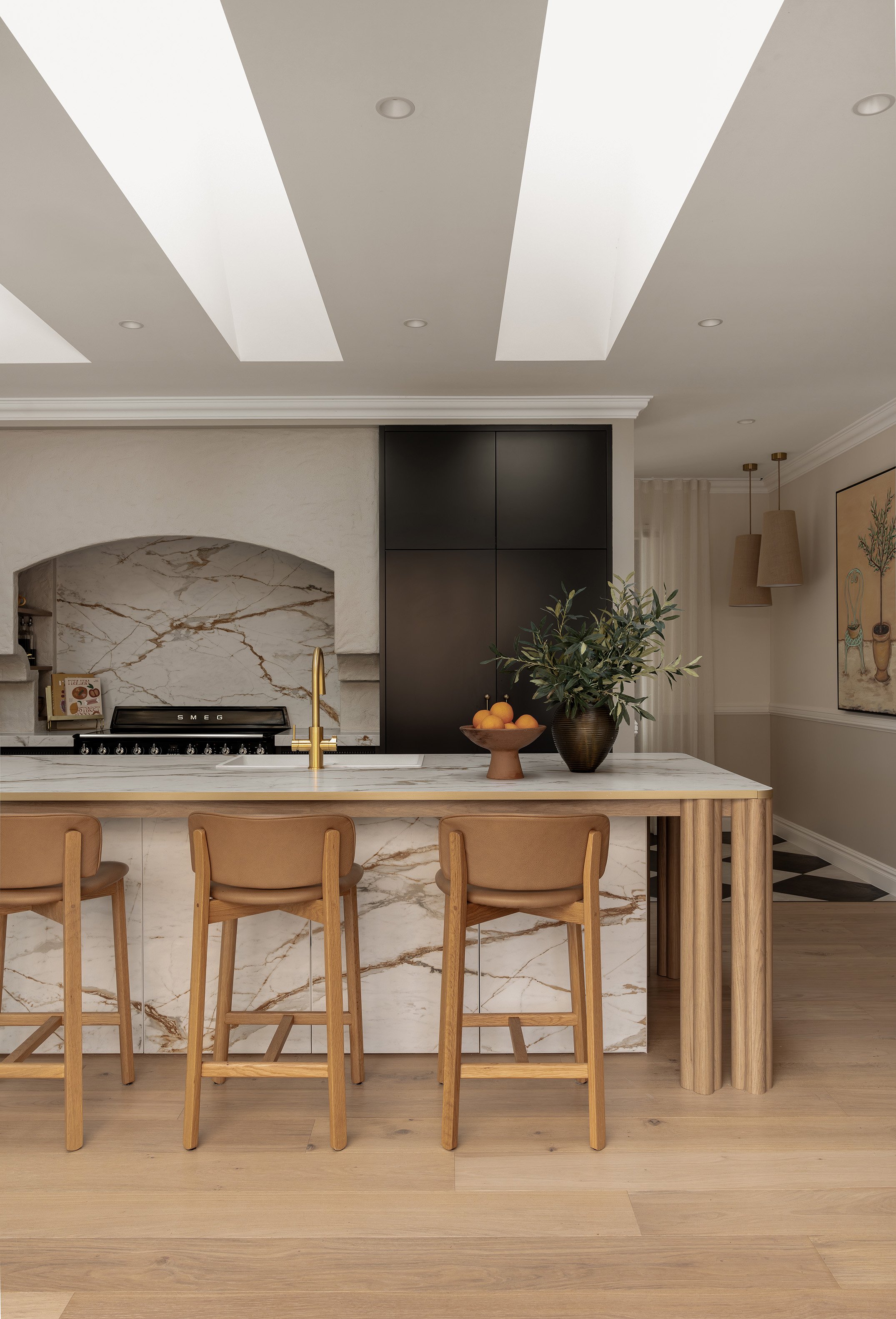 Modern kitchen with marble island, wooden bar stools, potted plant, and marble backsplash, with a stove and black cabinet in the background.