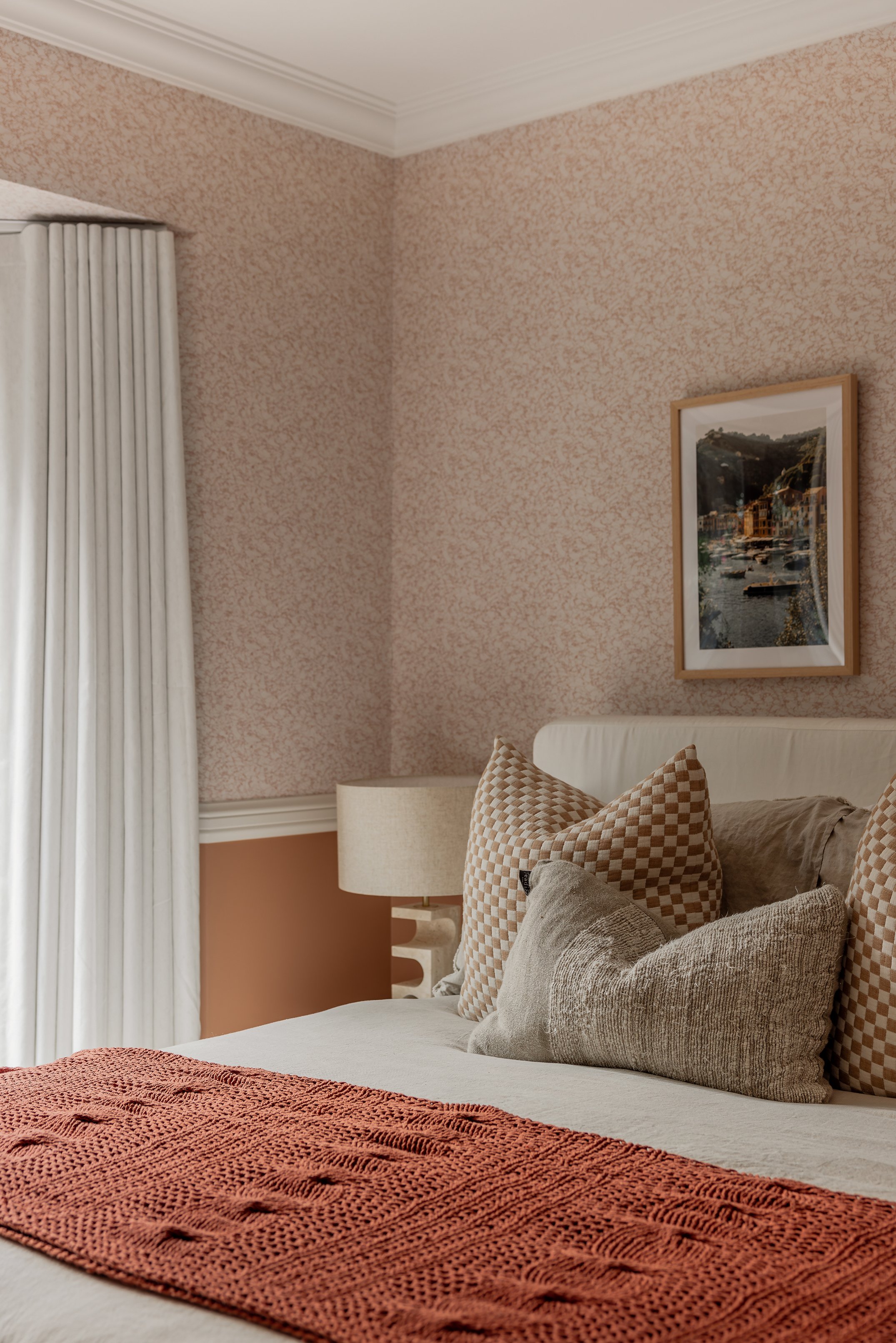 A cozy bedroom corner featuring a bed with beige and patterned pillows, an orange knit blanket, a bedside lamp, a framed landscape picture on a pink textured wall, and cream-colored curtains.