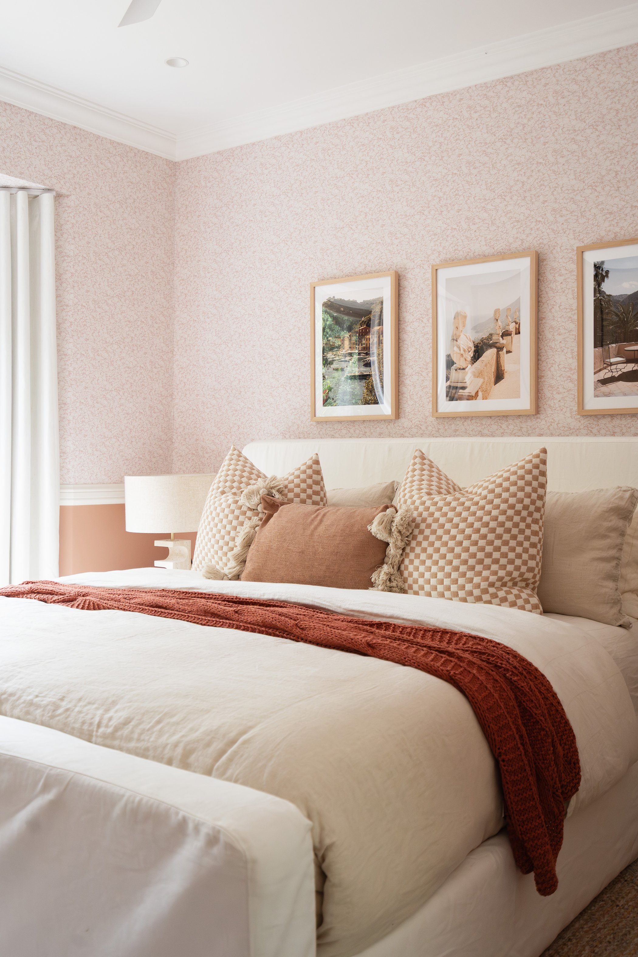 Cozy bedroom with a white bed, beige and checkered pillows, a red throw blanket, pink patterned walls, framed landscape pictures, and a white curtain.