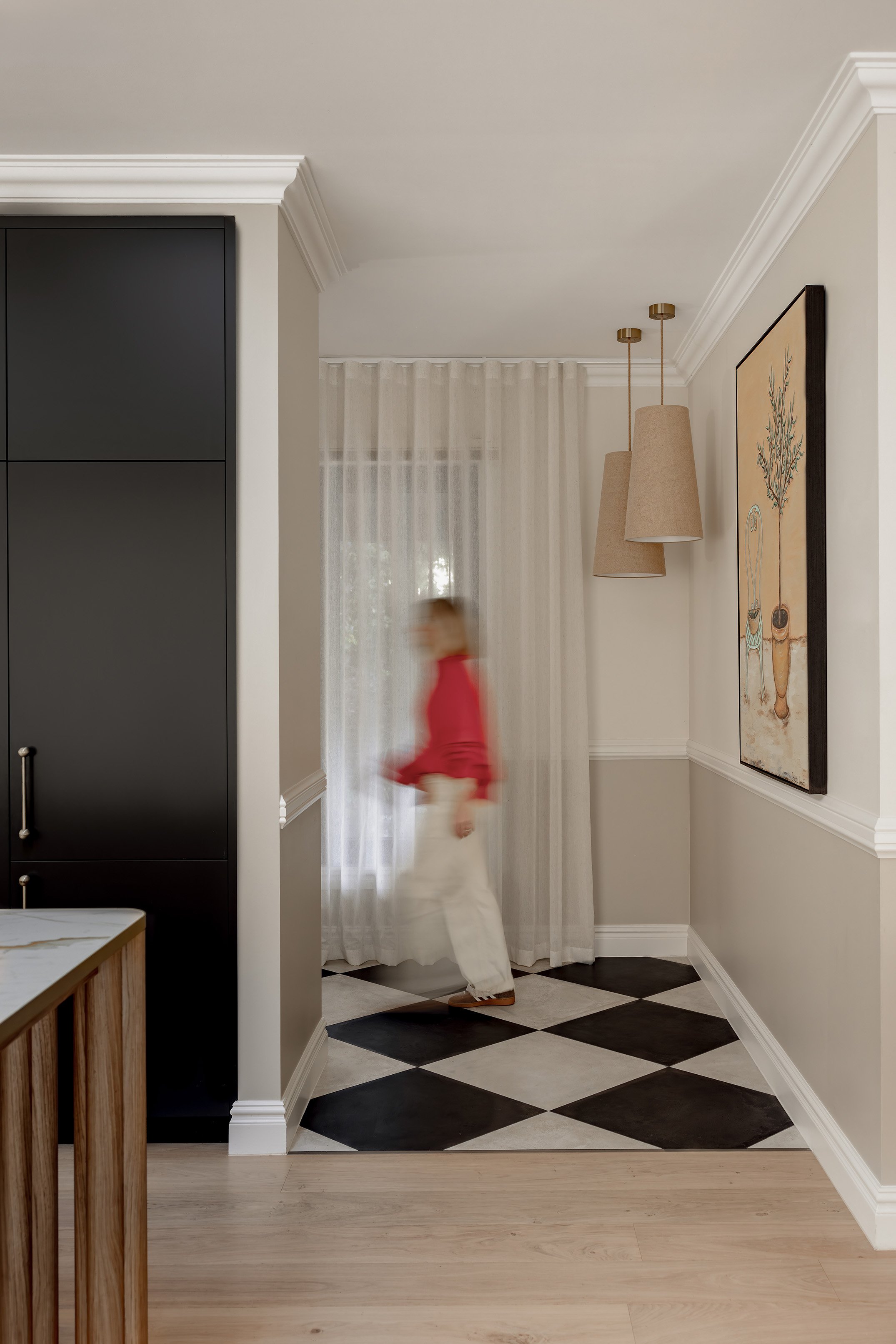 A woman in a red top and white pants walking in a home with black and white checkered flooring, beige walls, and hanging beige pendant lights.