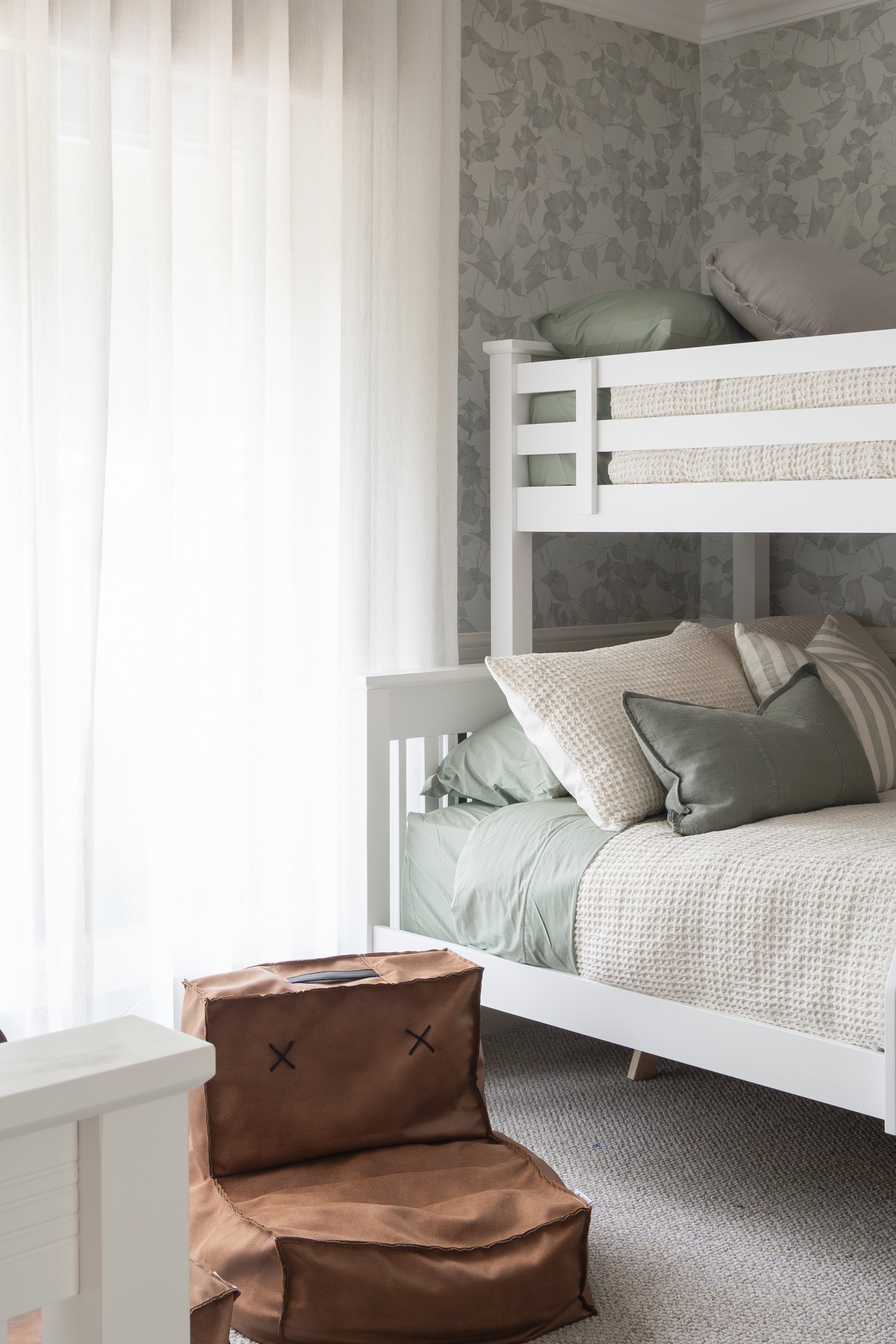 Bedroom with white bunk beds, green and beige bedding, patterned wallpaper, and a brown beanbag chair on a beige carpet.