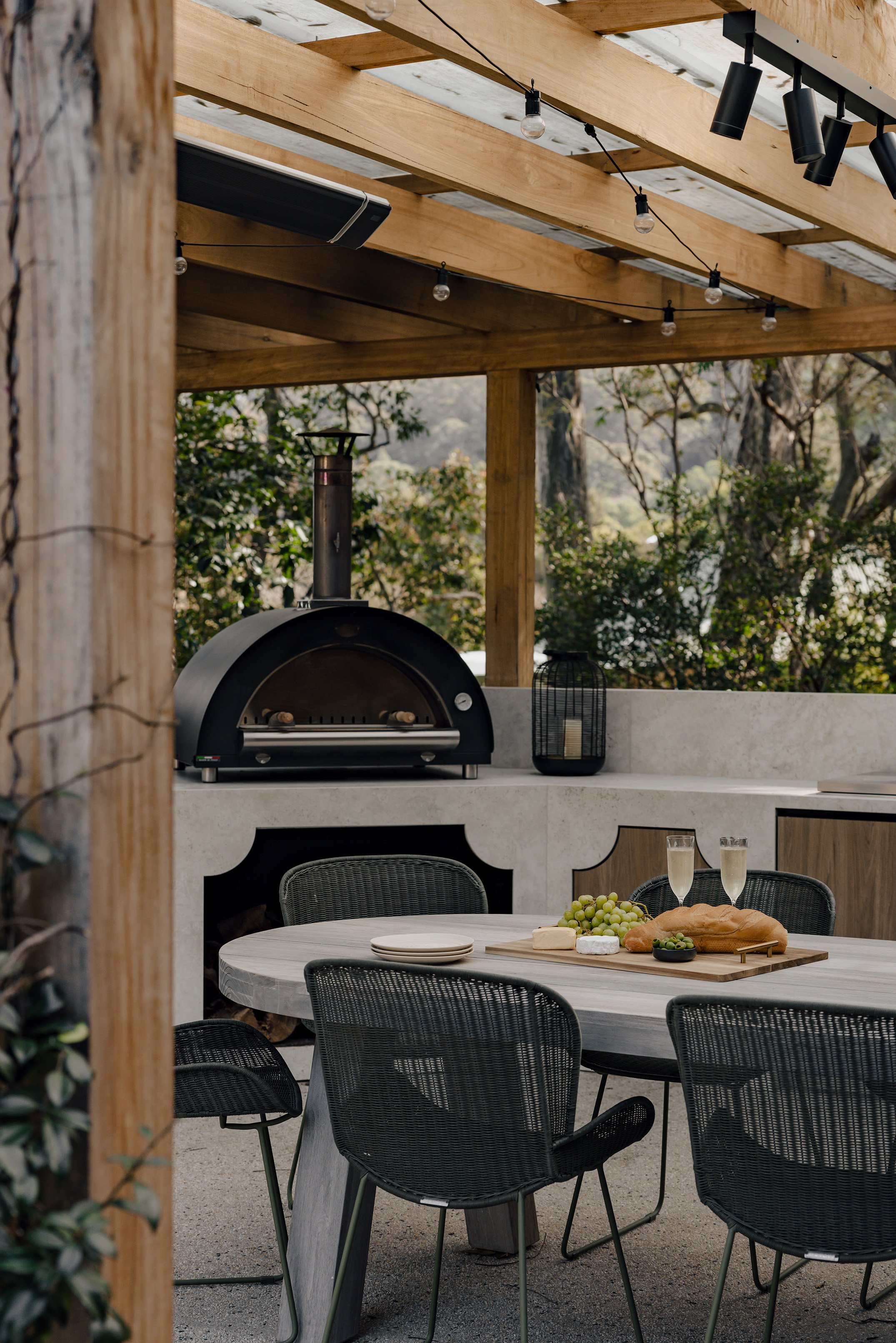 Outdoor dining area with a round table set with cheese, grapes, bread, and glasses of champagne. Behind the table is an outdoor pizza oven, with a lantern and trees in the background under a wooden pergola with string lights.