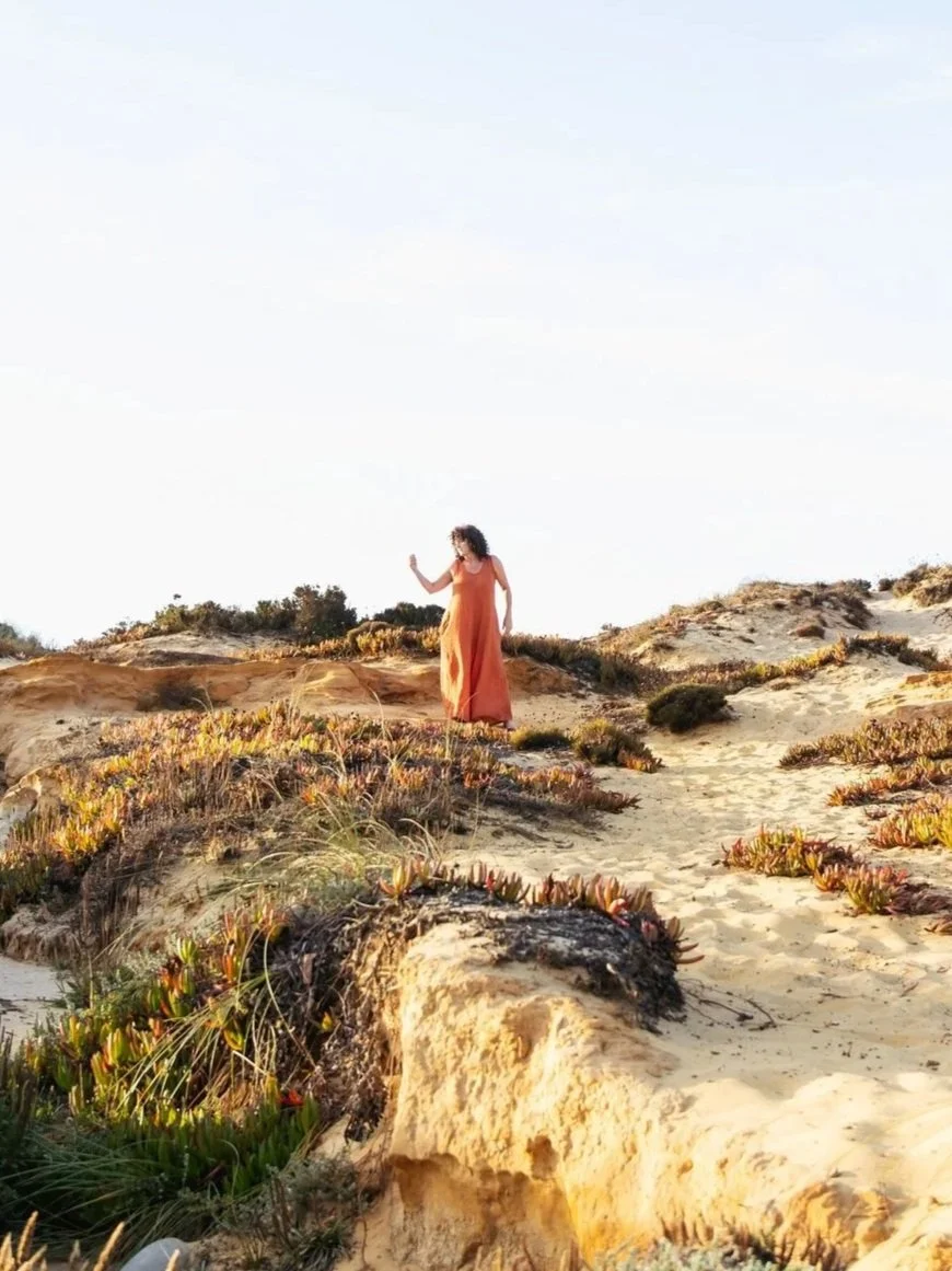 A woman in an orange dress walking on sandy terrain with sparse vegetation, under a light blue sky.