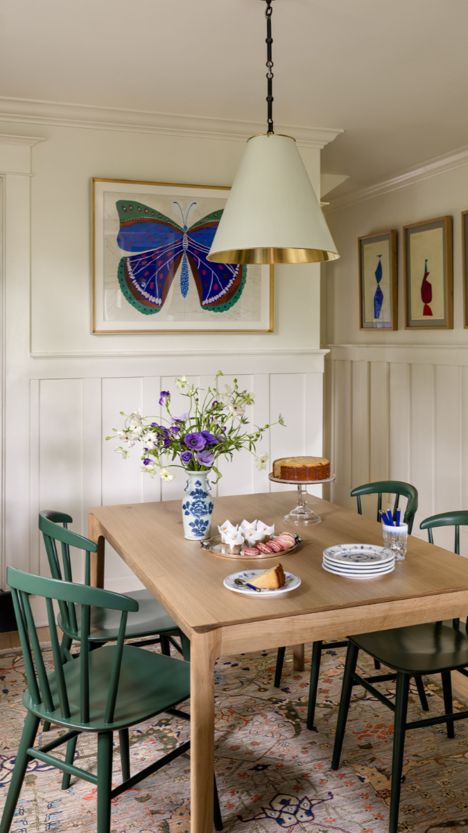 This image showcases a bright dining nook featuring a light wood table paired with classic spindle-back chairs painted in a green. The space is enhanced by white wainscoting walls, a neutral color palette, and warm natural textures, creating a clean 