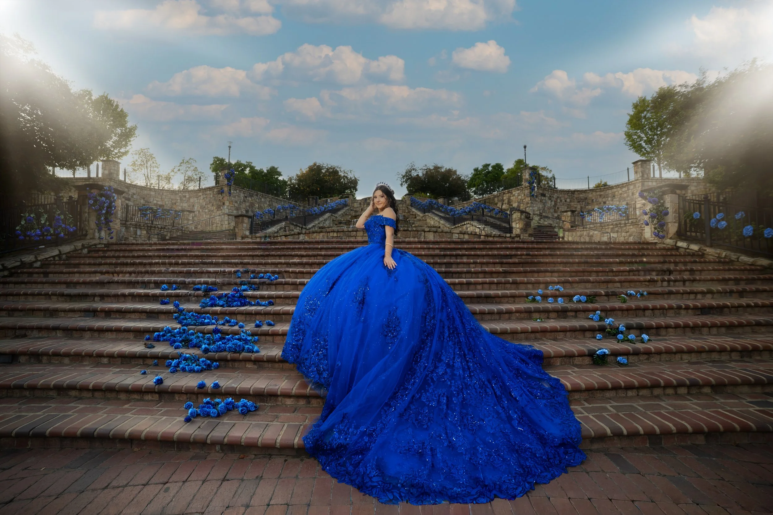 A woman in a royal blue ball gown standing on a set of brick stairs decorated with blue roses outdoors under a partly cloudy sky.