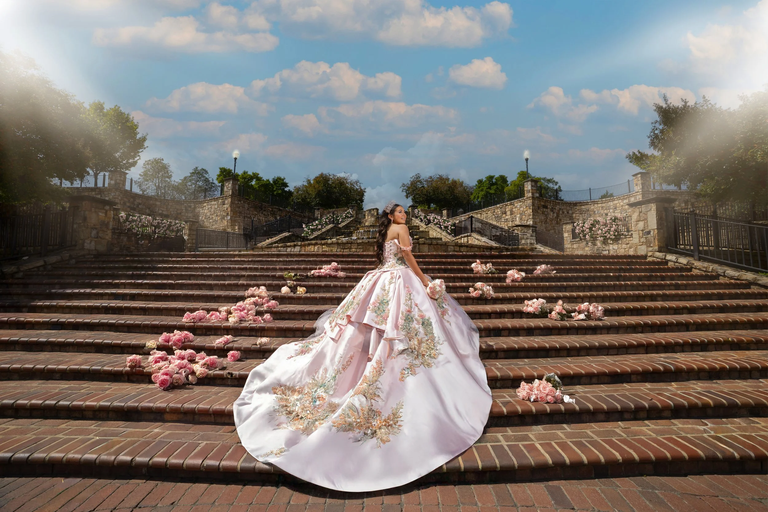 A woman in a pink floral wedding gown sitting on brick stairs scattered with pink roses, outdoors on a sunny day with trees and a stone wall in the background.
