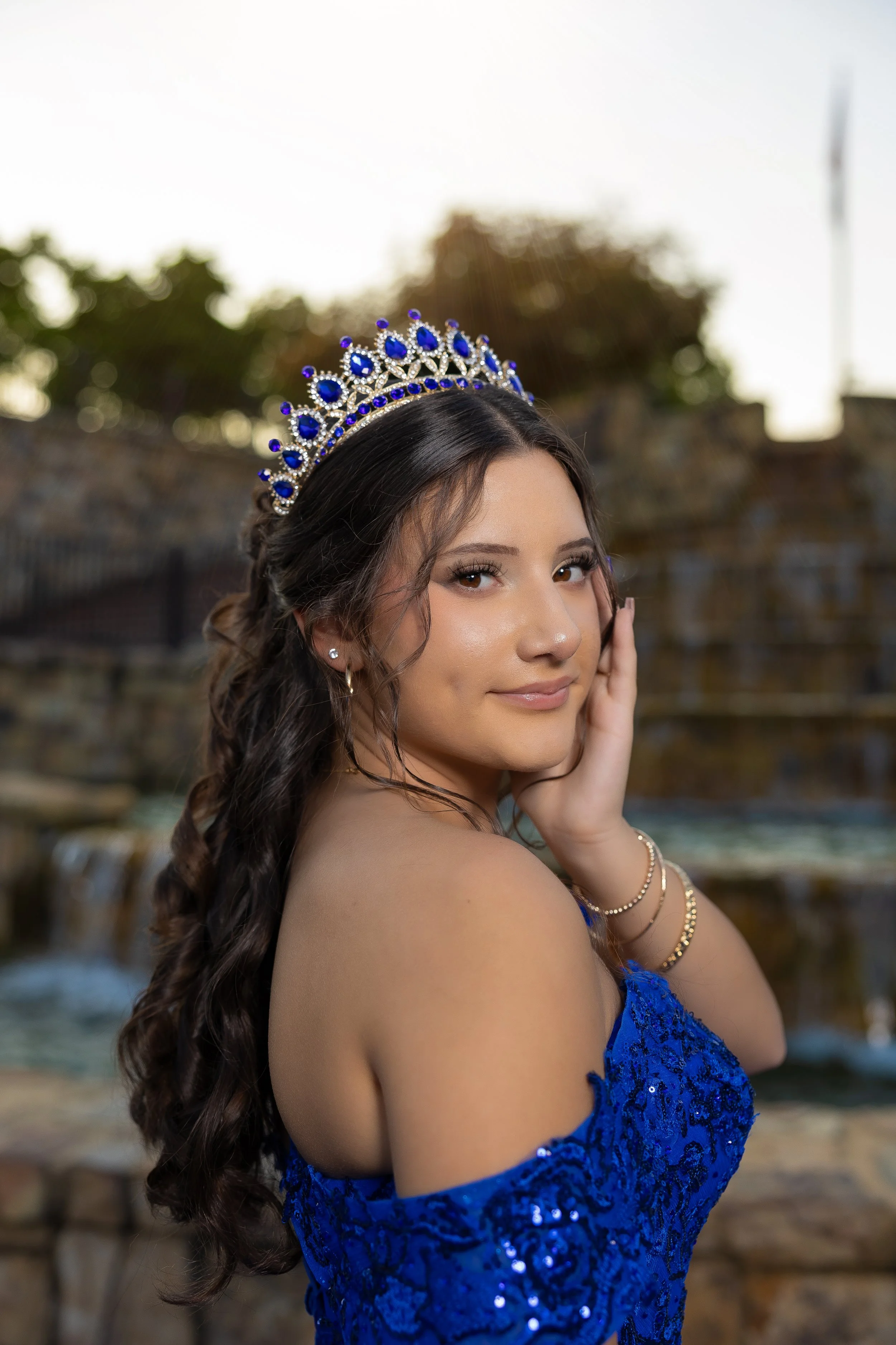 A young woman with long dark hair wearing a blue dress and a crown, posing outdoors with a blurred background of a waterfall and trees.