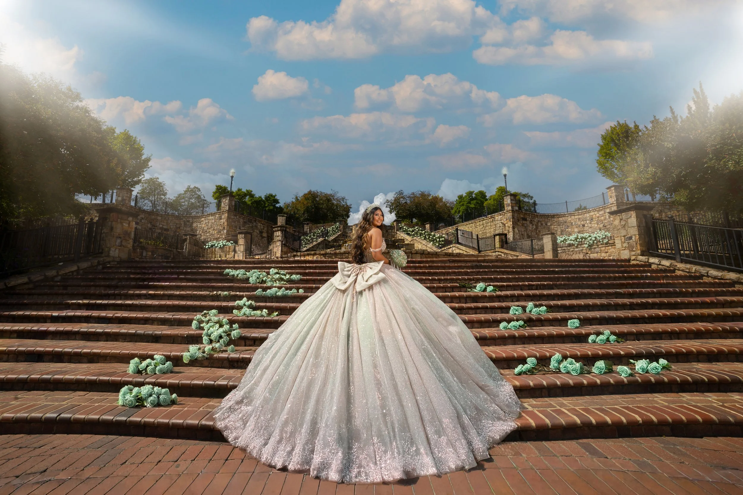 A woman in a white wedding gown with a big bow in the back stands on a wide brick staircase decorated with scattered pale turquoise roses. The backdrop features stone walls, green trees, and a partly cloudy sky.