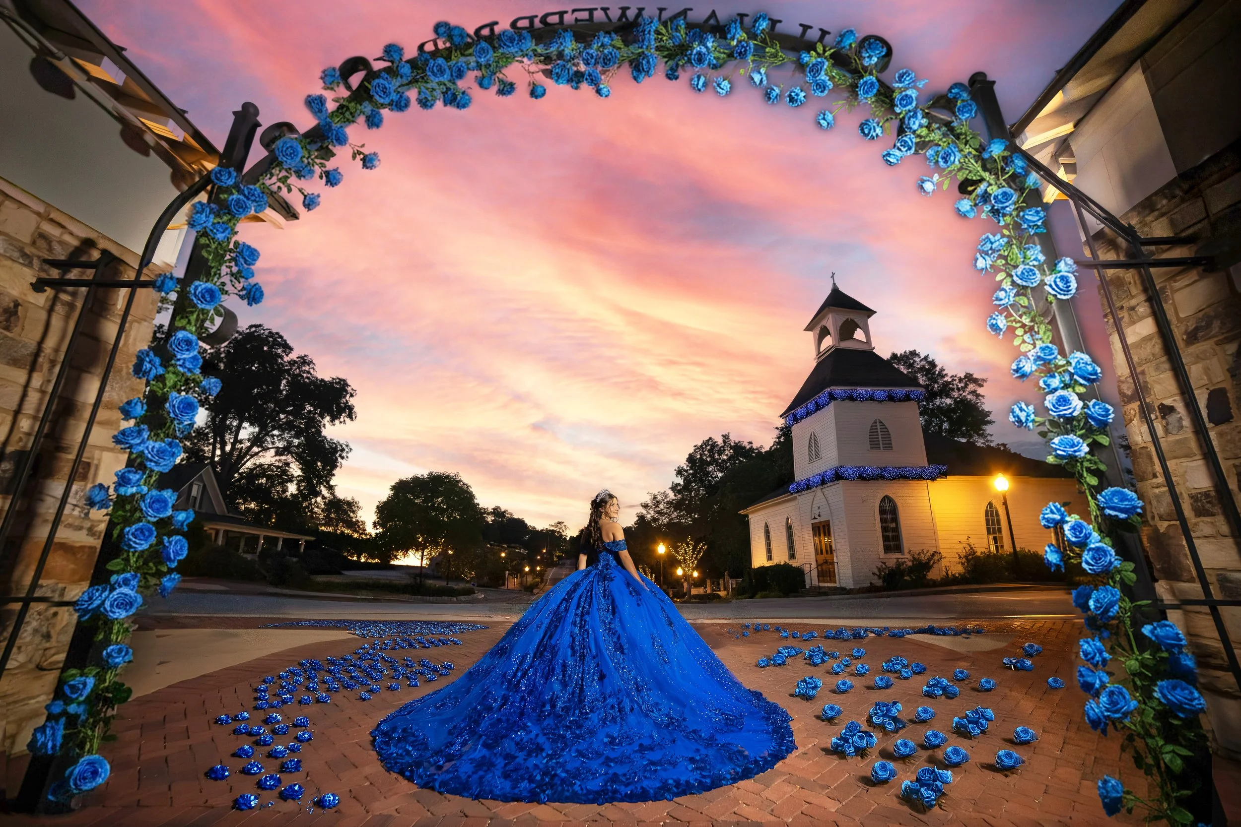 A woman in a blue dress standing in front of a church at sunset, with an arch decorated with blue roses and scattered rose petals on the brick ground.