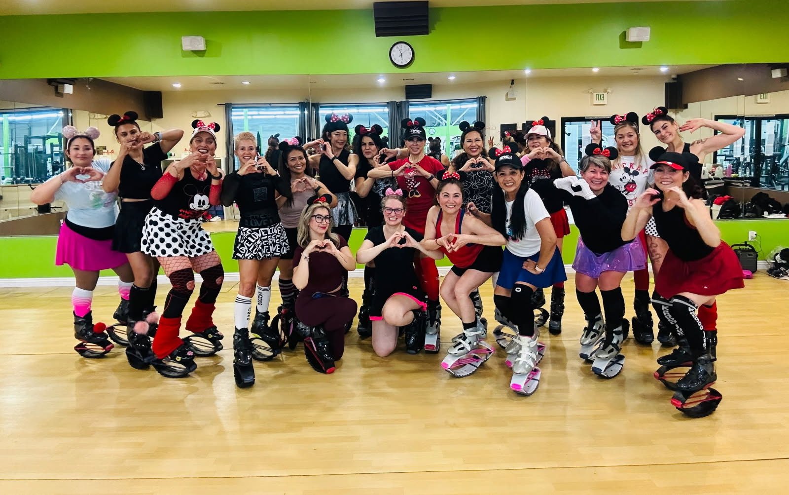 A group of people wearing kangaroo jump boots and Mickey Mouse ears, posing in a gym with a green wall and wooden floor.
